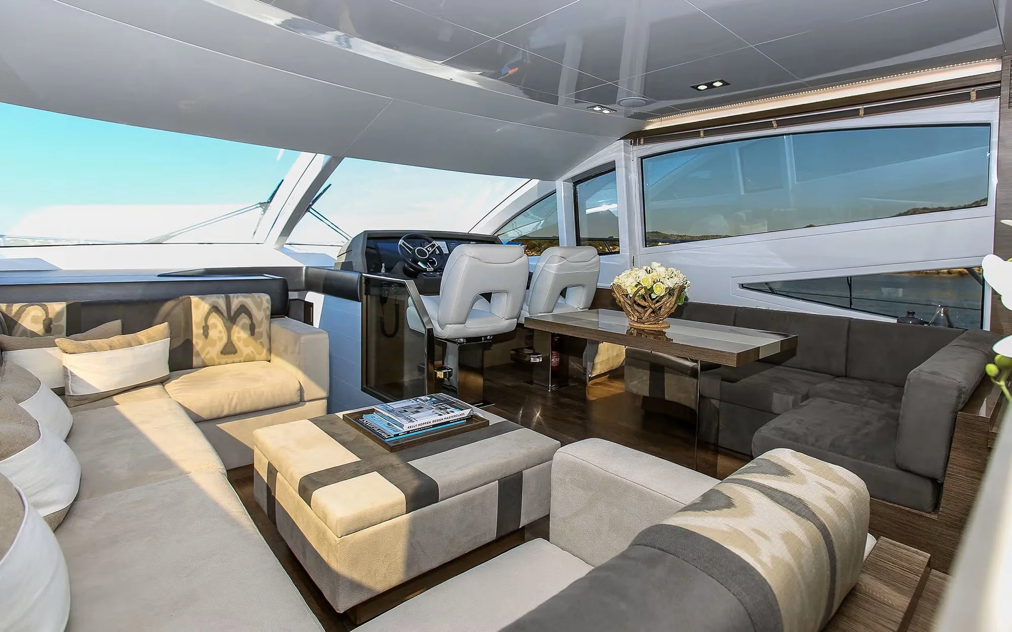Luxury yacht interior with a steering wheel, white leather captain chairs, a wooden table with a bouquet of white flowers, beige and dark gray sofas, and large windows showing water and sky outside.