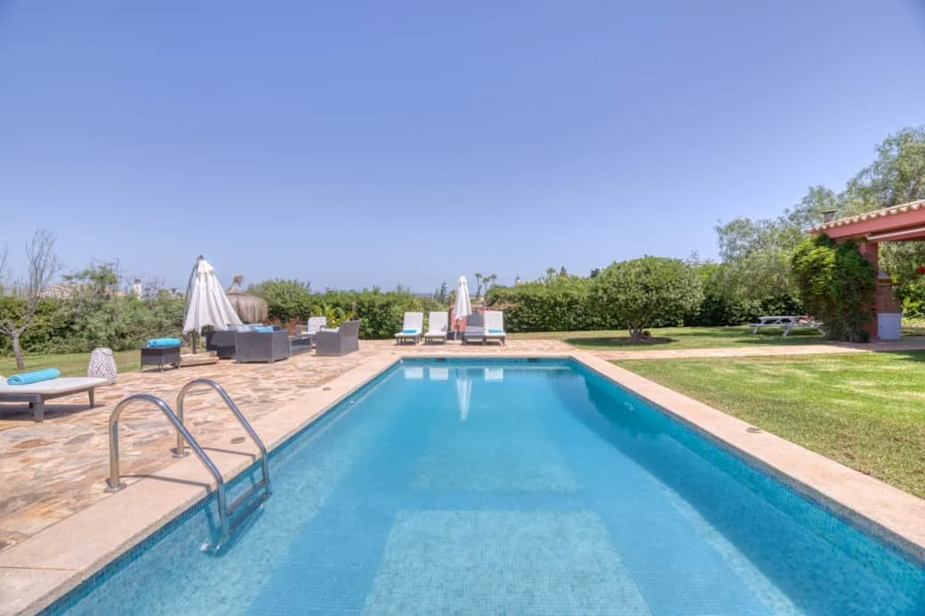 An outdoor swimming pool with lounge chairs and umbrellas on a sunny day, surrounded by green trees and a grassy area.