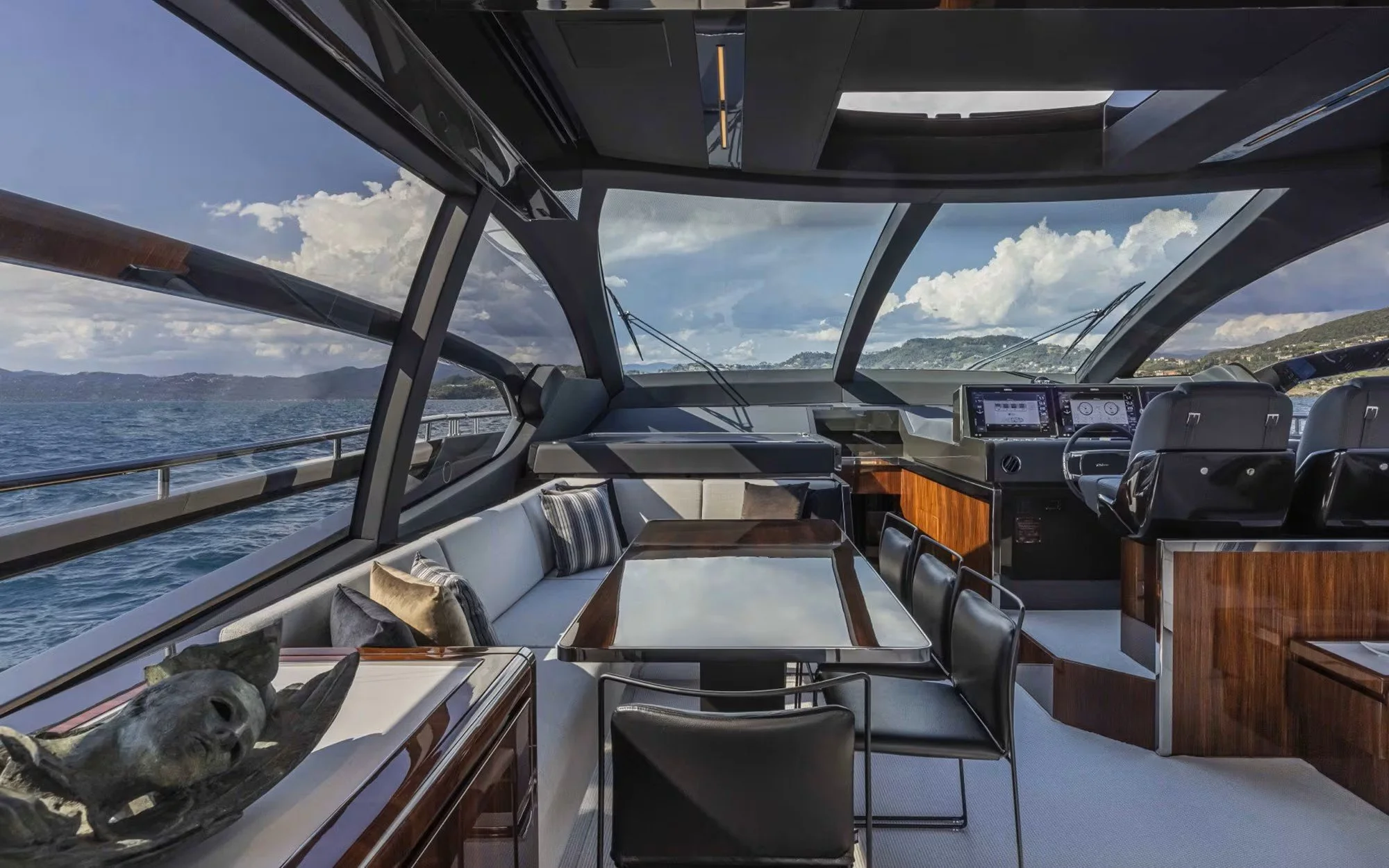 Interior view of a yacht's living and helm area with a dining table, chairs, plush sofa with pillows, wood paneling, and navigation screens, overlooking water and hills outside through large windows.