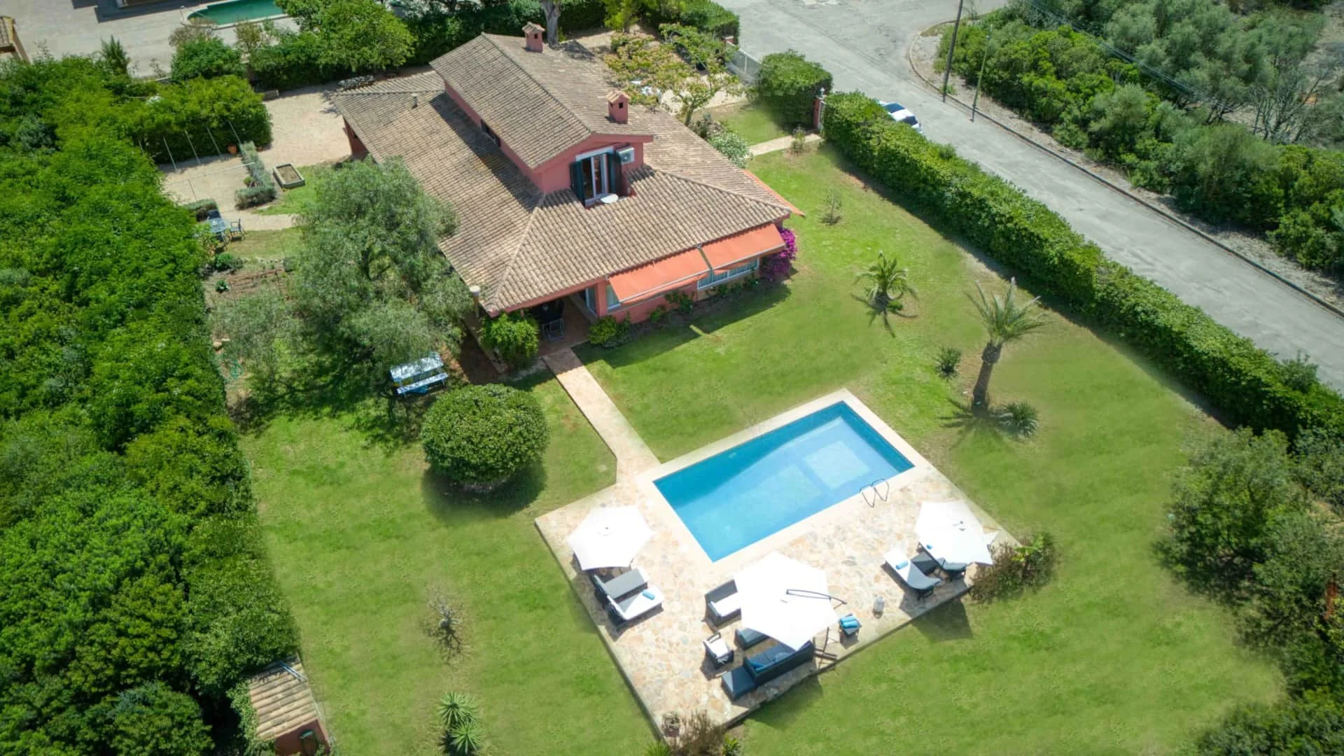 Aerial view of a house with a red-tiled roof, surrounded by green trees and bushes, featuring a swimming pool with lounge chairs and umbrellas in the backyard.