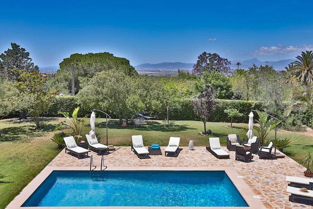 A backyard with a swimming pool, lounge chairs, umbrellas, and outdoor seating area surrounded by trees and greenery with mountains in the distance.