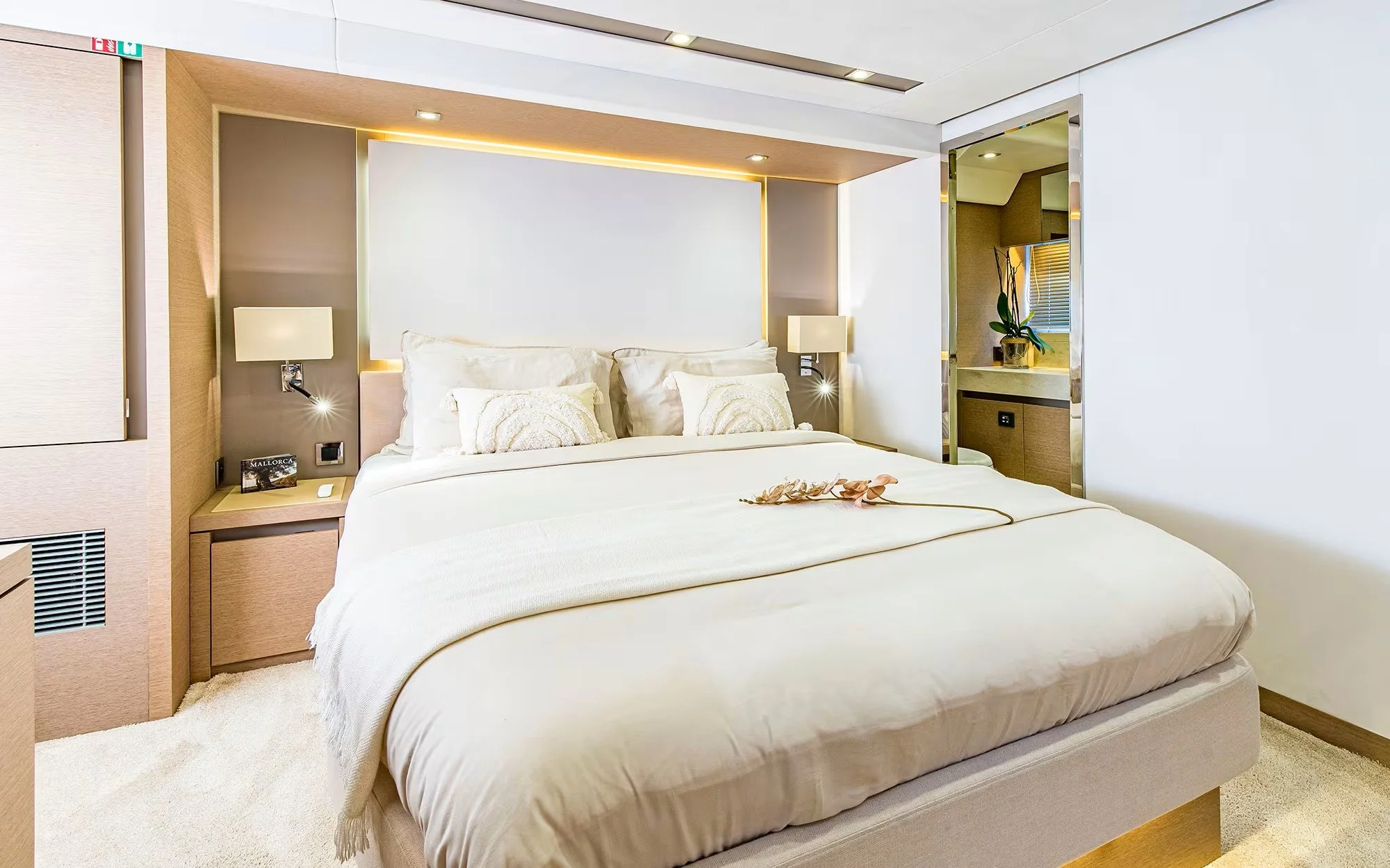 A modern hotel bedroom with a large bed, white linens, and decorative pillows. There are two wall-mounted lamps, a nightstand with a small book and tissue box, and a mirror reflecting a window with blinds. The room features a neutral color palette wi