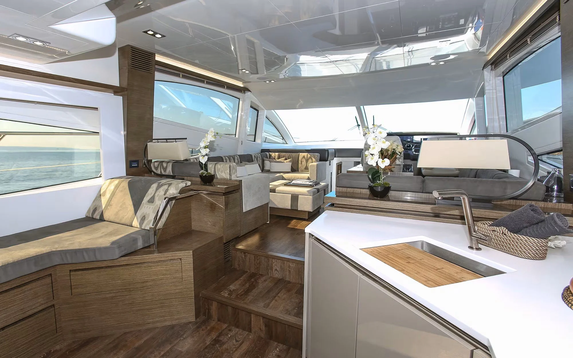 Interior of a yacht with large windows, a seating area with beige and gray cushions, wooden steps, and a kitchenette with a white countertop, sink, and drying rack. Decorative white orchids are on the counter and near the seating area.