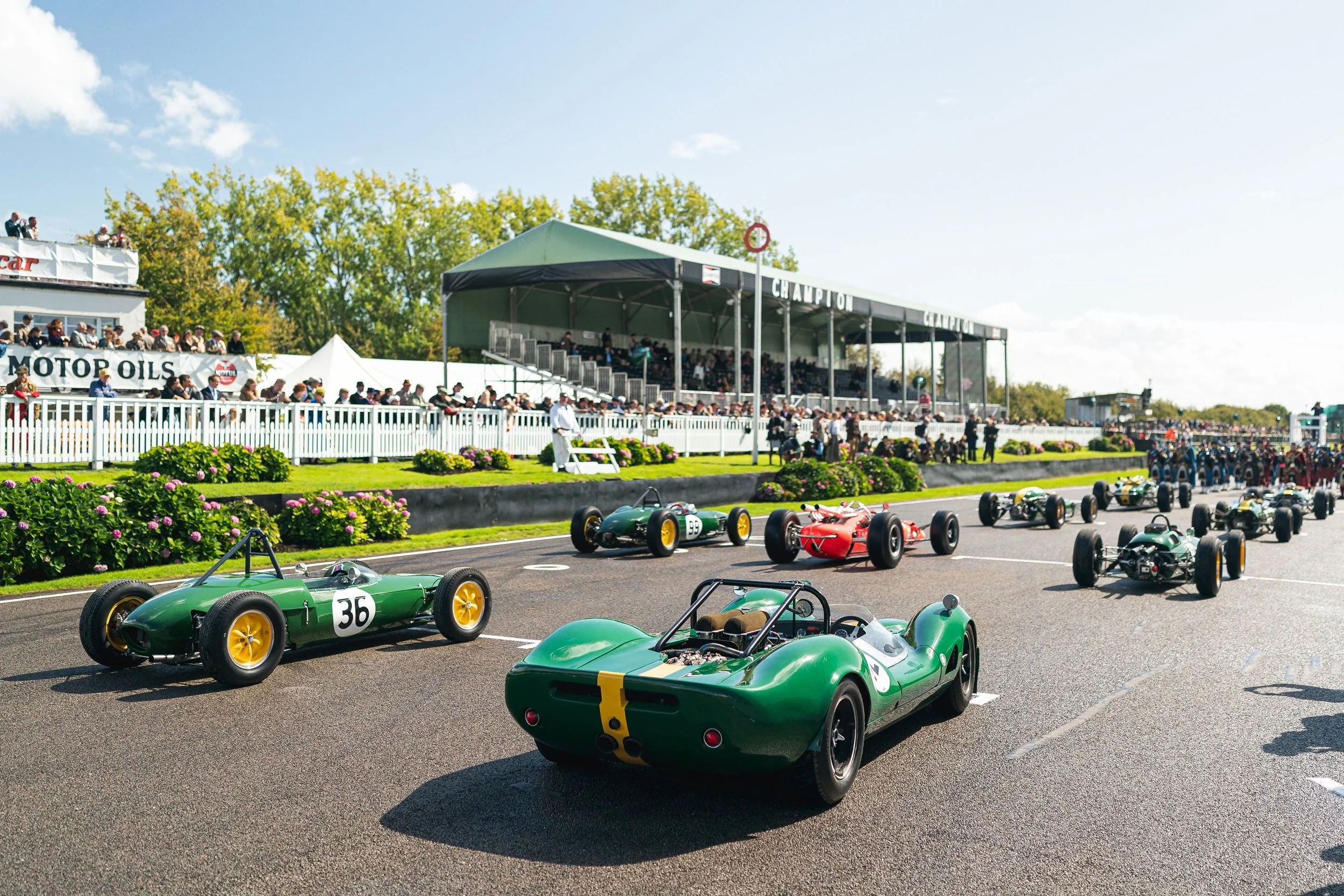 Vintage race cars lined up on starting grid at a race track, with spectators and a grandstand in the background, lush greenery and blue skies above.