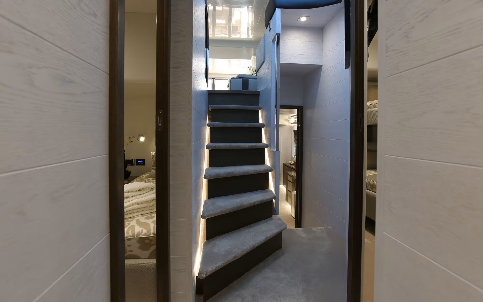 A modern staircase with gray carpeted steps and LED strip lighting along the side. The staircase is situated in a narrow corridor with light-colored wood-paneled walls, leading up to a second floor. There is a glimpse of a bedroom to the left and a s