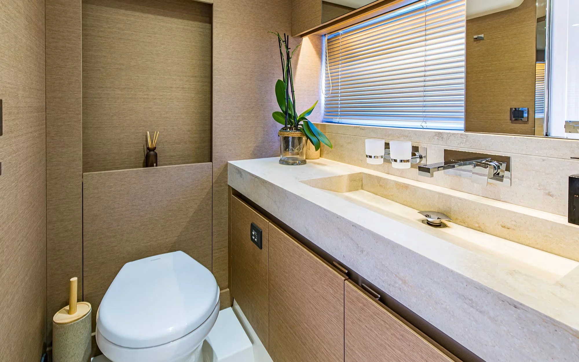 A modern bathroom with beige walls, a white toilet, a beige countertop with a built-in sink, a potted plant, a window with blinds, and minimal decor.