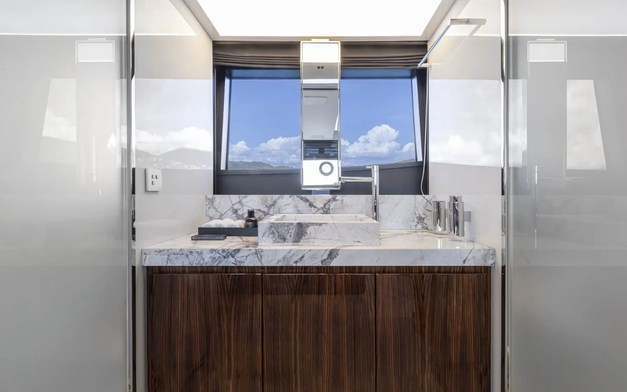 Modern bathroom with marble sink, wooden vanity, large window showing a blue sky with clouds.