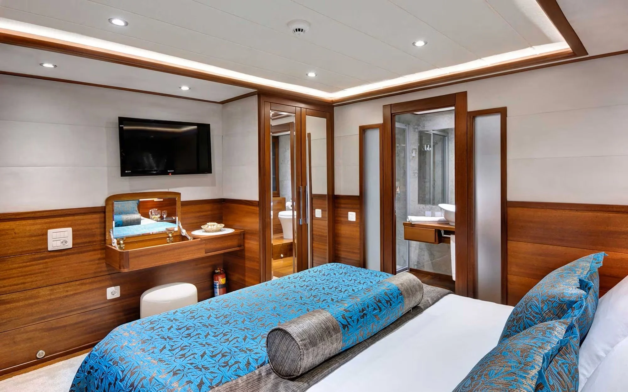 Hotel room with a flat-screen TV on the wall, a bed with blue patterned bedspread and cushions, and a wooden partition leading to a bathroom with a glass shower.