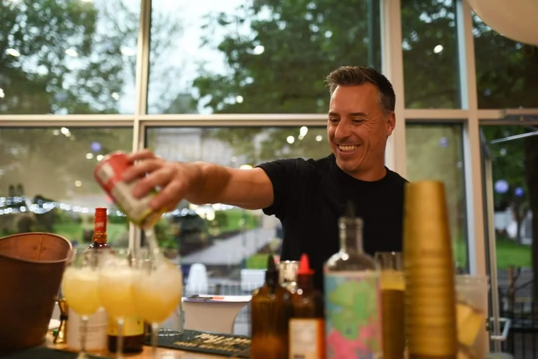 Un mixologue souriant prépare un cocktail ou une boisson derrière un bar avec diverses bouteilles et ingrédients, avec une grande fenêtre donnant sur un espace extérieur vert.