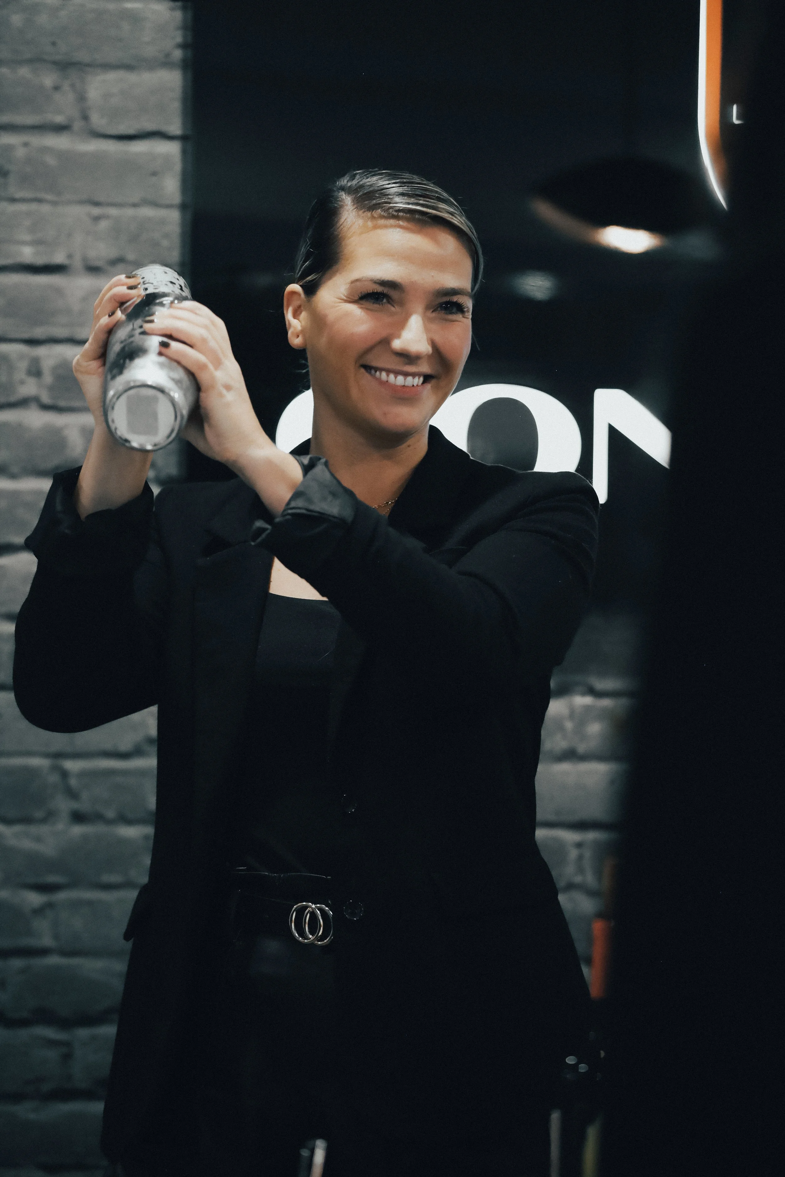 Une mixologue souriante shake un cocktail dans un événement corporatif, arrière-plan en pierre gris et enseigne lumineuse.