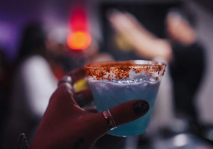 Main tenant un verre de cocktail bleu avec tajin sur le rebord, dans un bar ou une fête.