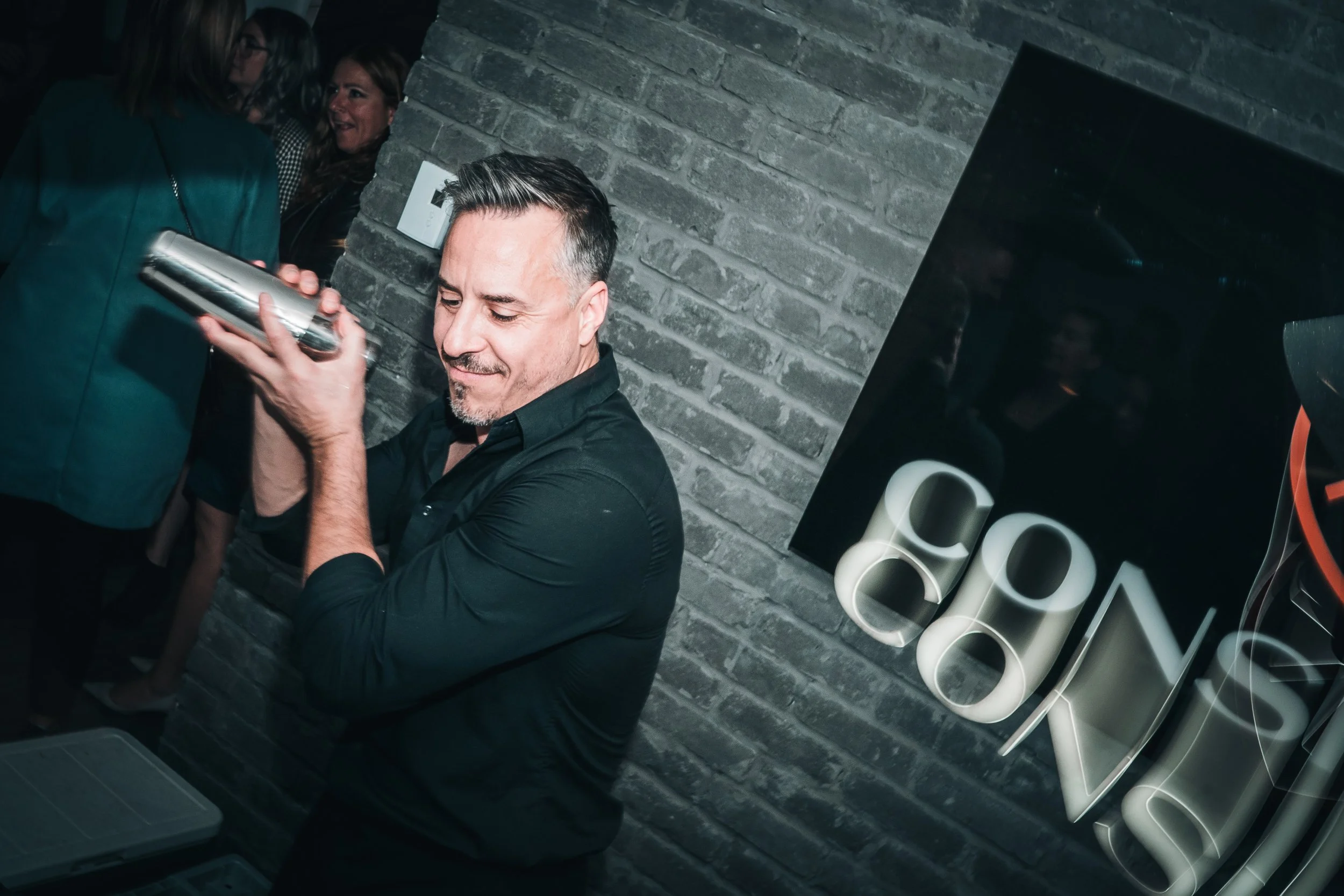 Un mixologue préparant un cocktail lors d'une fête ou d'un événement, avec un groupe de personnes en arrière-plan et une enseigne lumineuse sur un mur en briques.