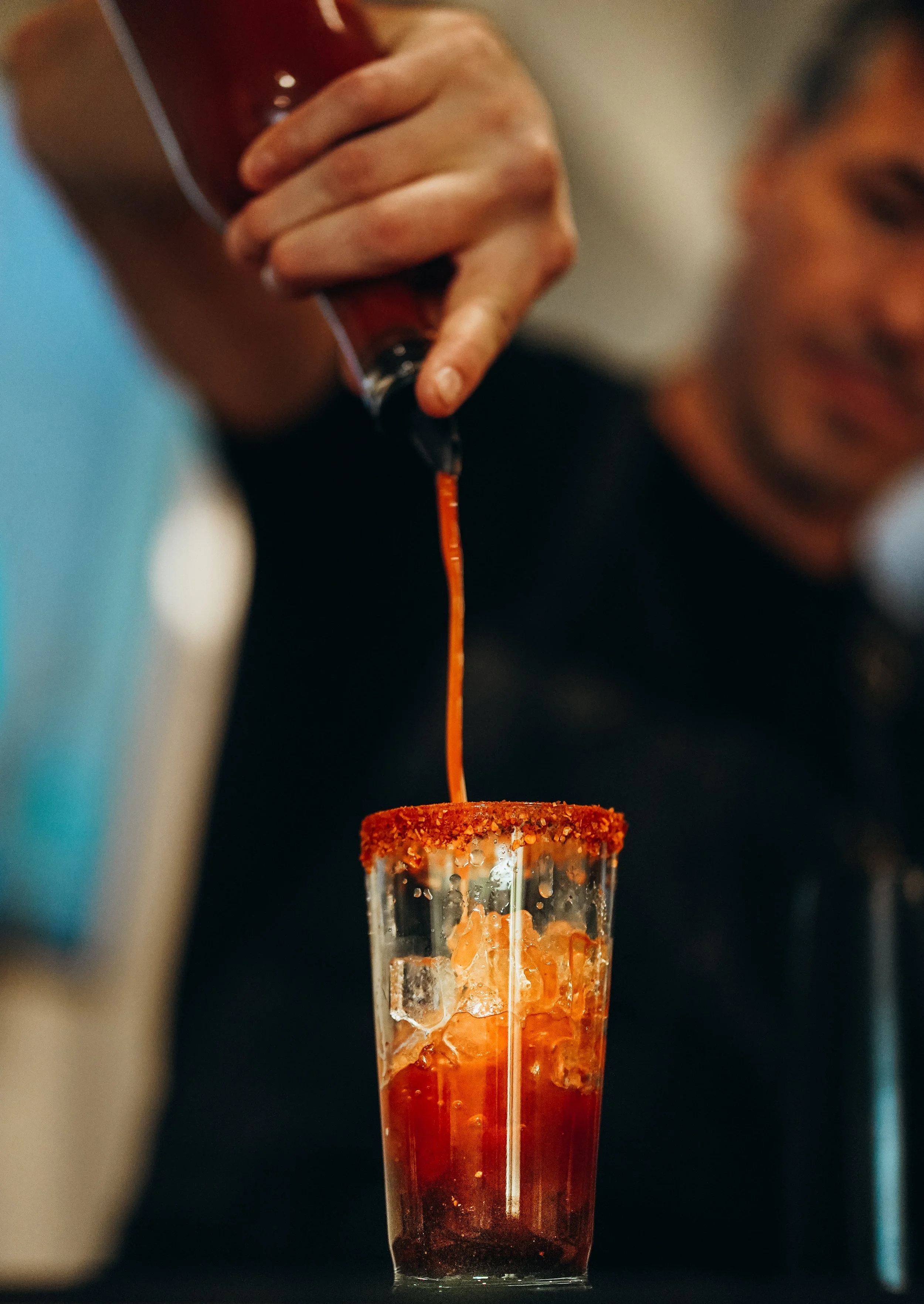 Une personne verse une sauce rouge épicée dans un verre contenant des glaçons, avec des bords givrés et un assaisonnement épicé au bord.