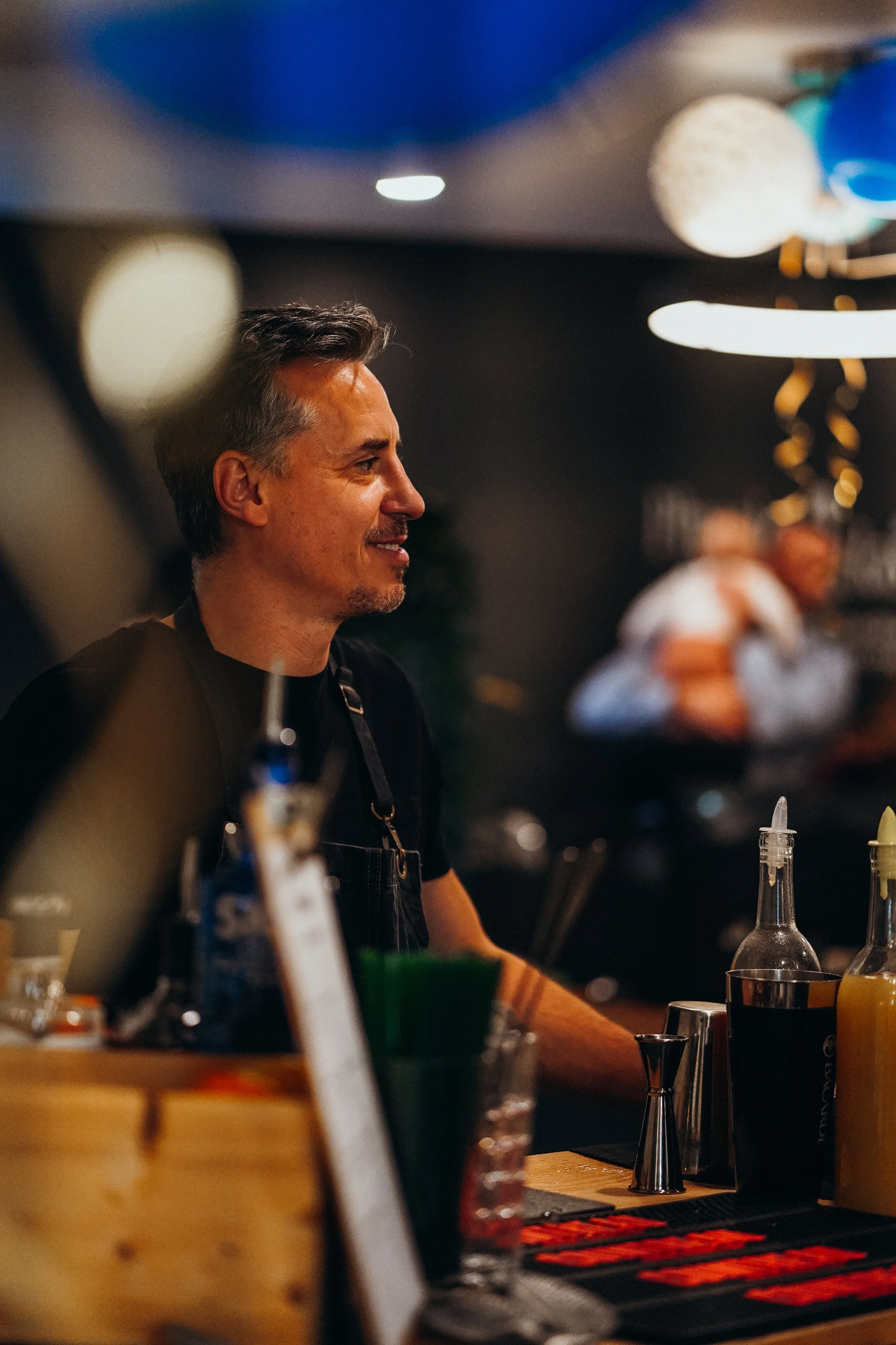 Homme barista souriant, en service derrière le comptoir d'un café ou bar, avec des bouteilles et accessoires en premier plan, décor moderne et ambiance chaleureuse.