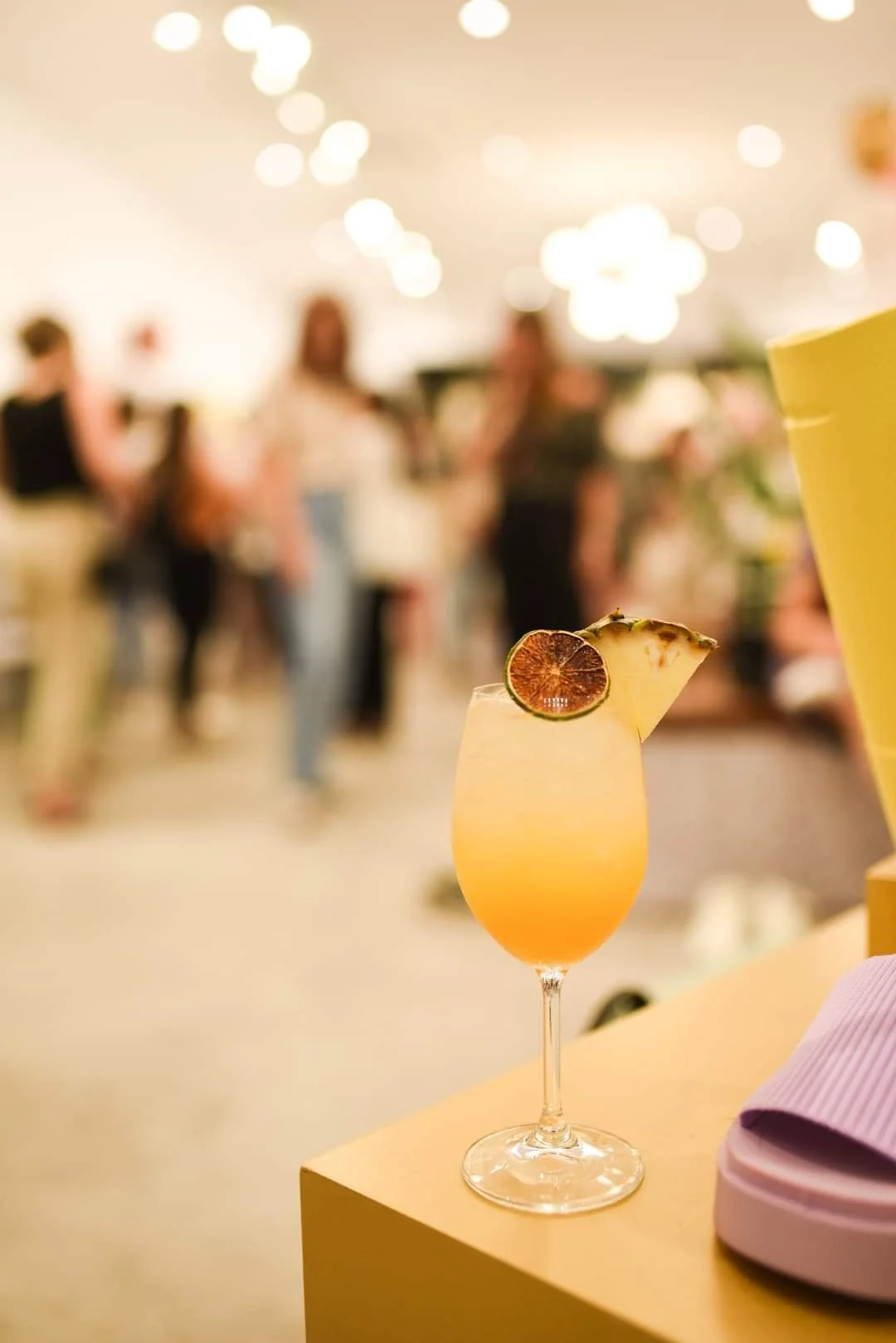 Un verre de cocktail orange décoré d'une tranche de citron vert, posé sur une table jaune, avec un fond flou de personnes dans une salle lumineuse.
