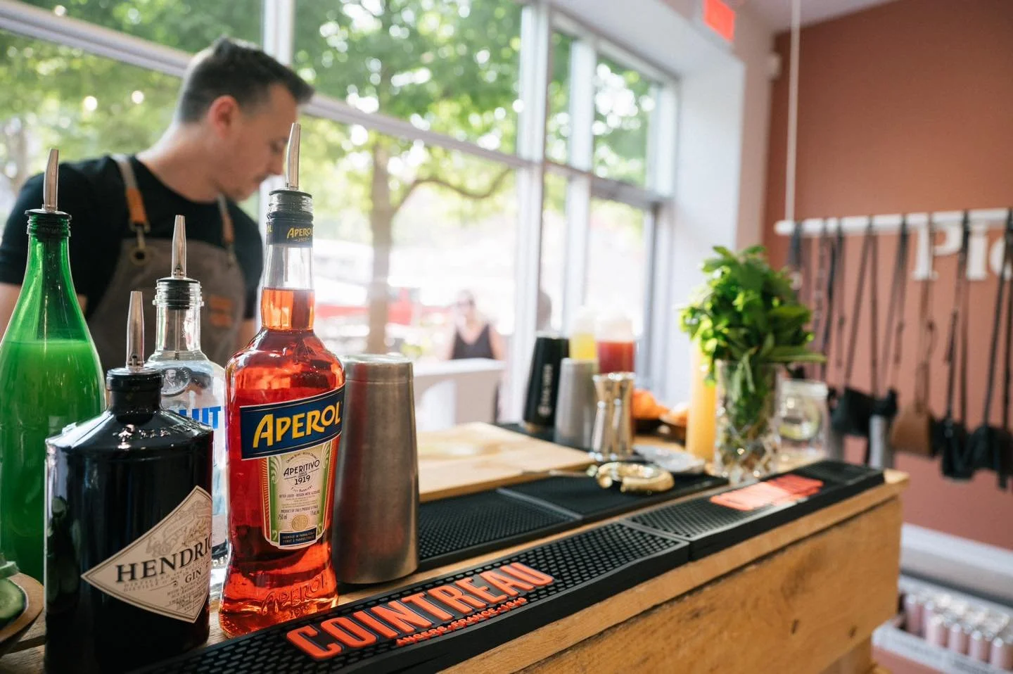 Bar avec diverses bouteilles d'alcool, un mixologue en arrière-plan, et un intérieur avec grande fenêtre laissant entrer la lumière naturelle, décor chaleureux et végétation visible dehors.