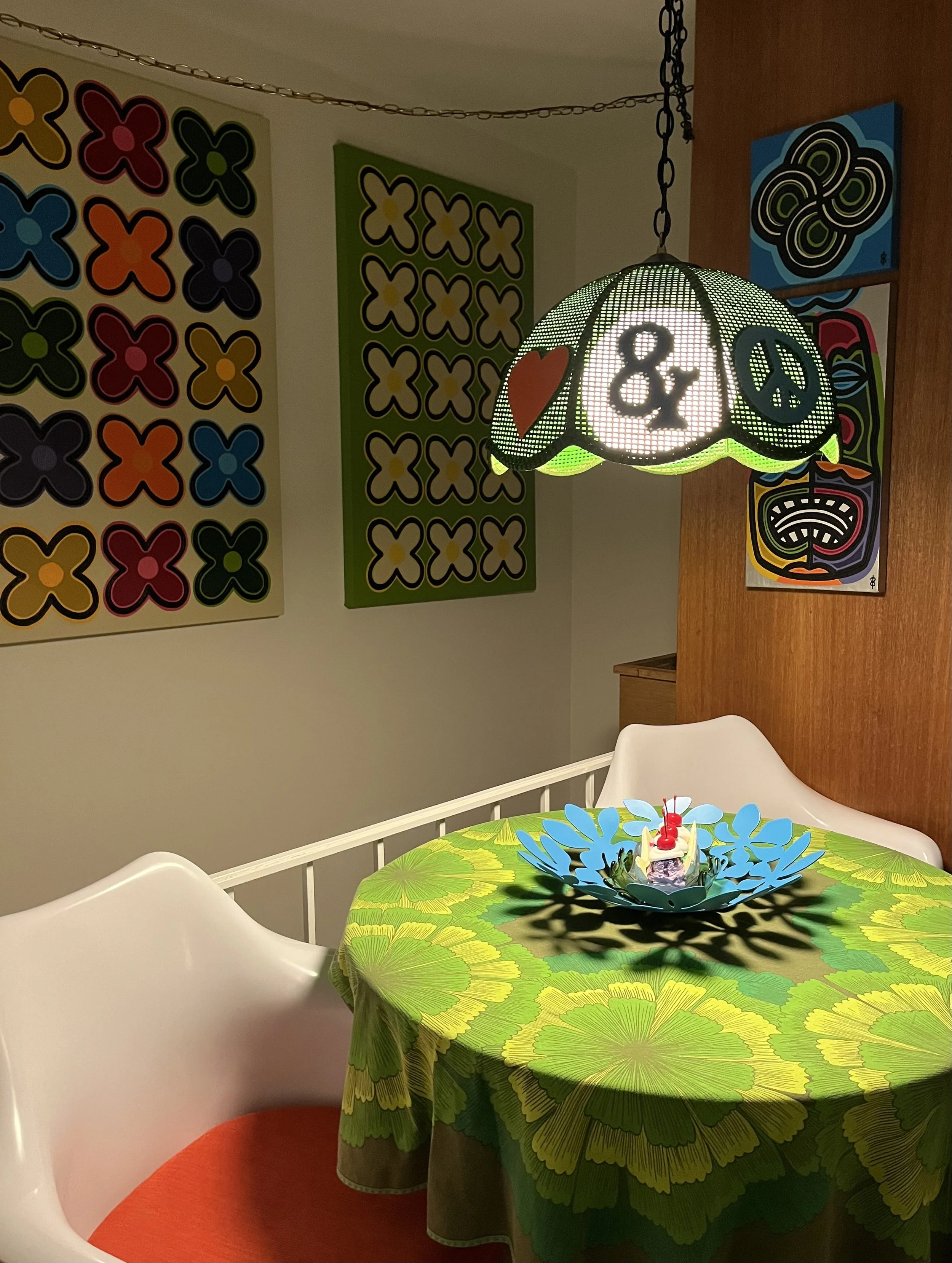 Front view of the hand-painted Peace & Love hanging light above a colorful table, showing the ampersand and peace-sign panels.