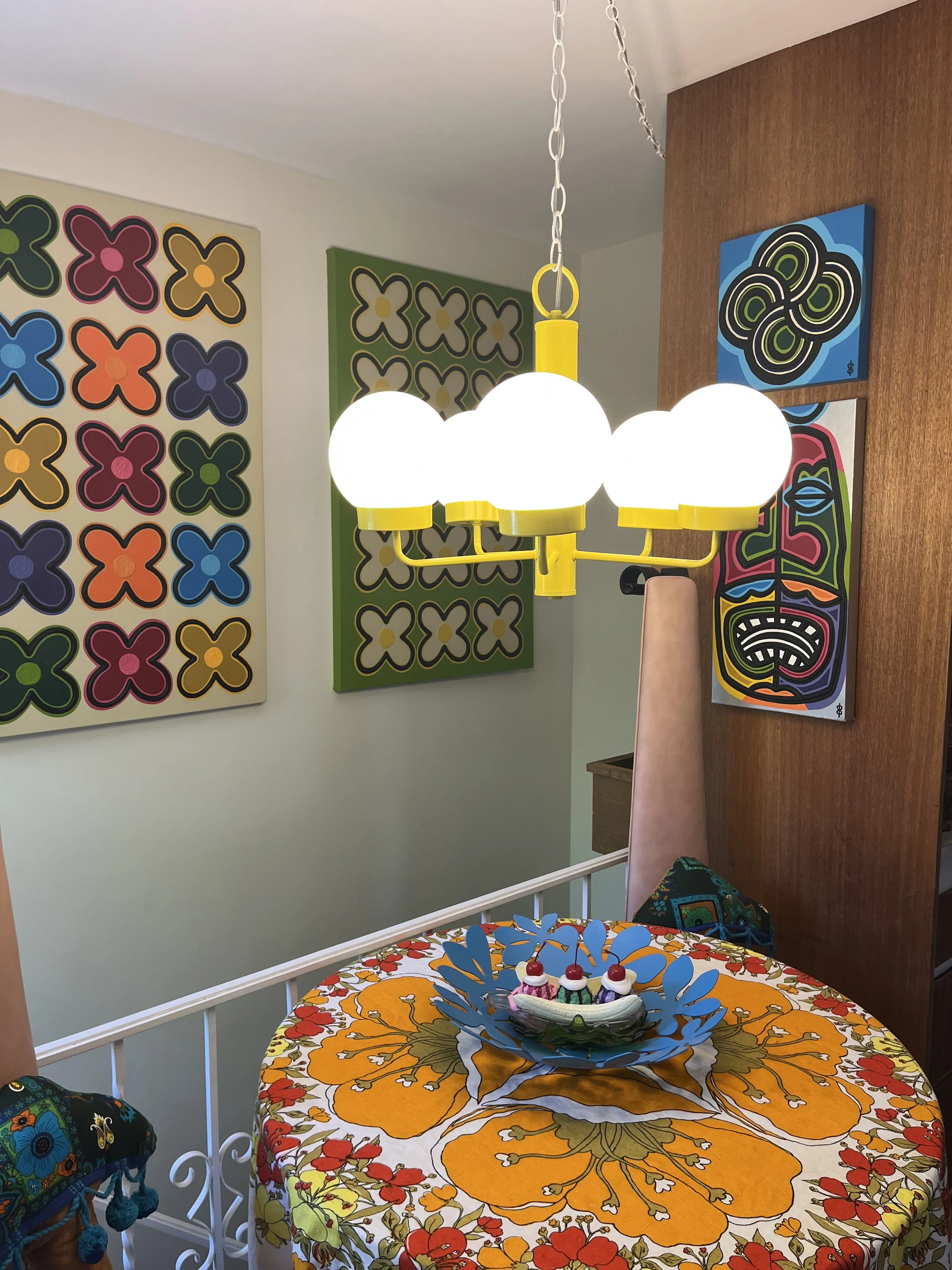Illuminated vintage canary yellow chandelier with five glowing white glass globes suspended above a retro floral dining table.
