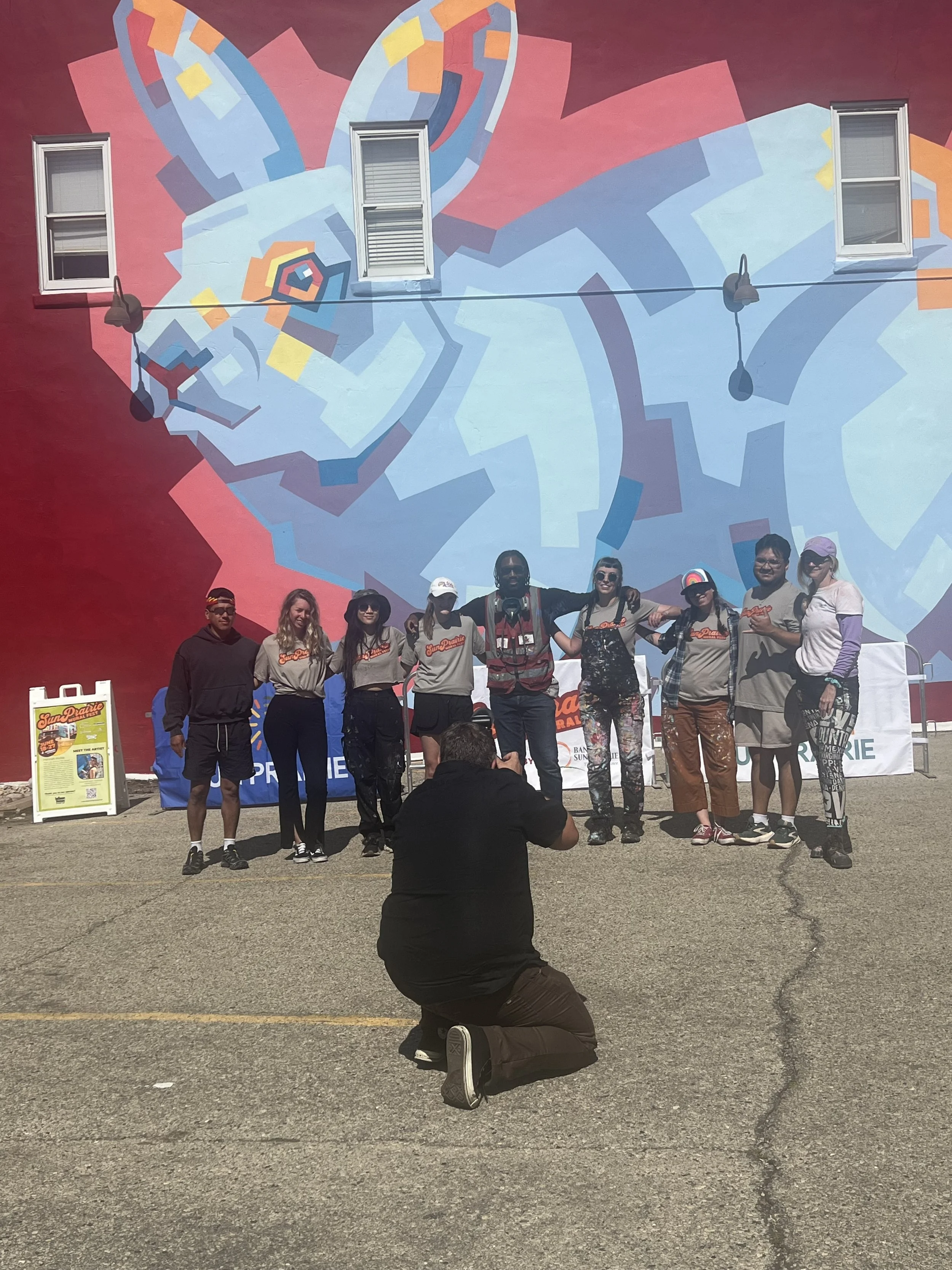 A group of people standing in front of a colorful mural of a chameleon painted on a wall. One person is kneeling in the foreground taking a photo. The group is smiling and posing for the picture.
