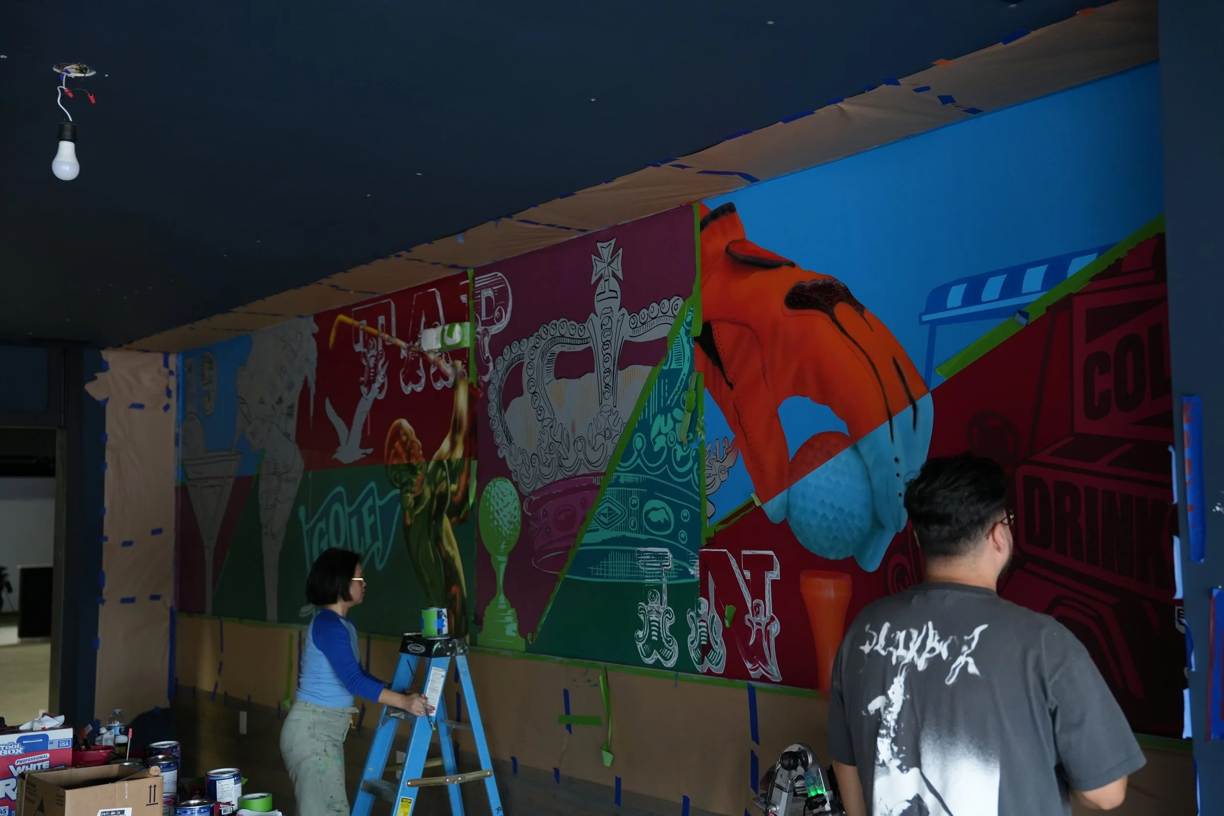 People working on painting a colorful mural on a large wall, including a woman on a ladder applying paint and a man with a roller. Painting supplies and open boxes are nearby.