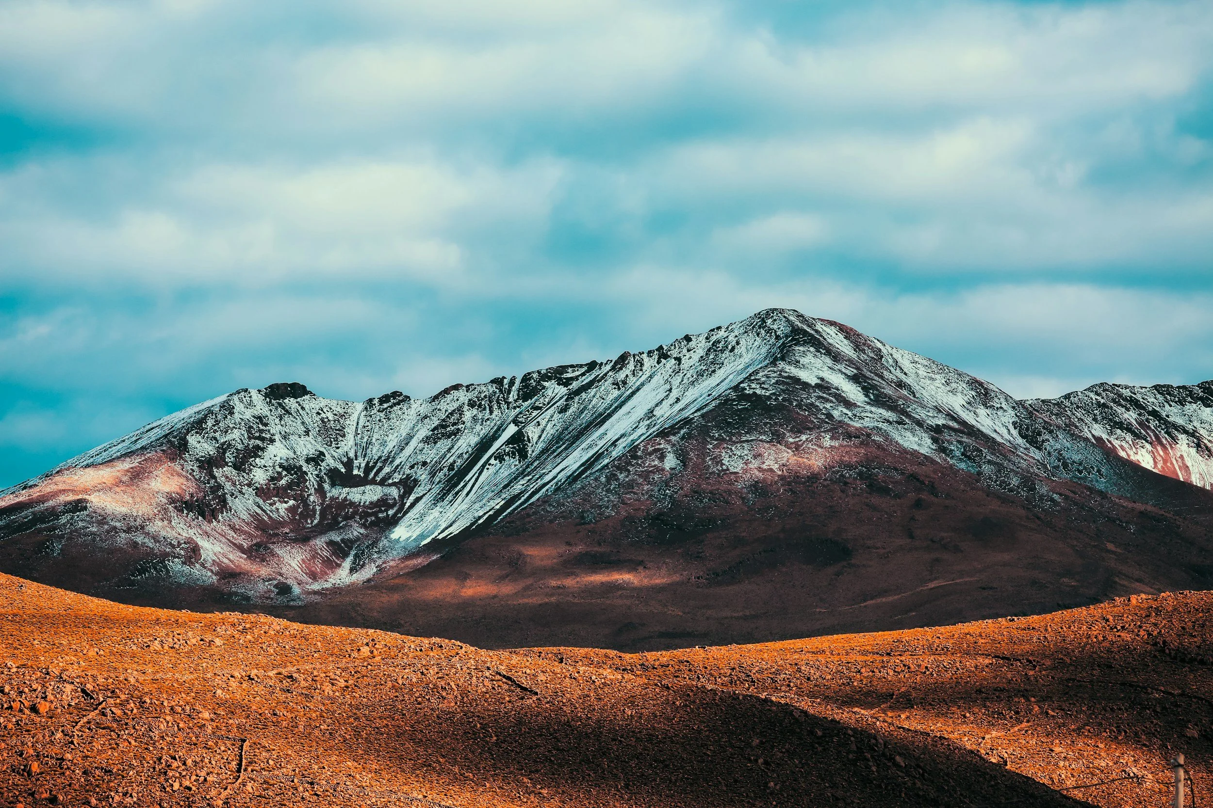 Snow-covered mountain with red and brown rocky terrain under a cloudy blue sky.