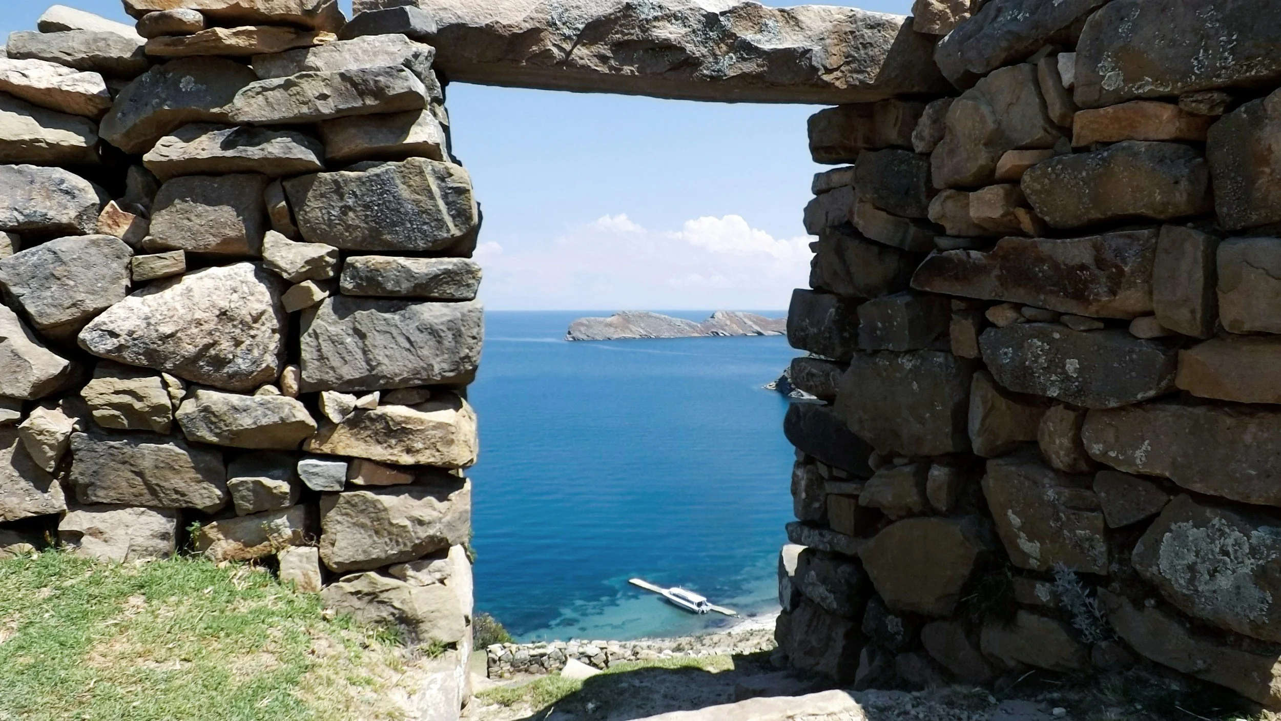 Stone wall with rectangular opening overlooking blue sea and small islands, with a boat at shore.