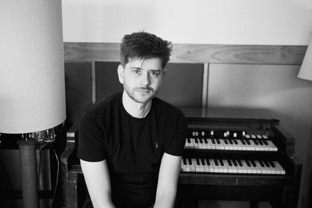 Black and white portrait of Oli Jacobs sitting in front of an organ with a wooden panel background.