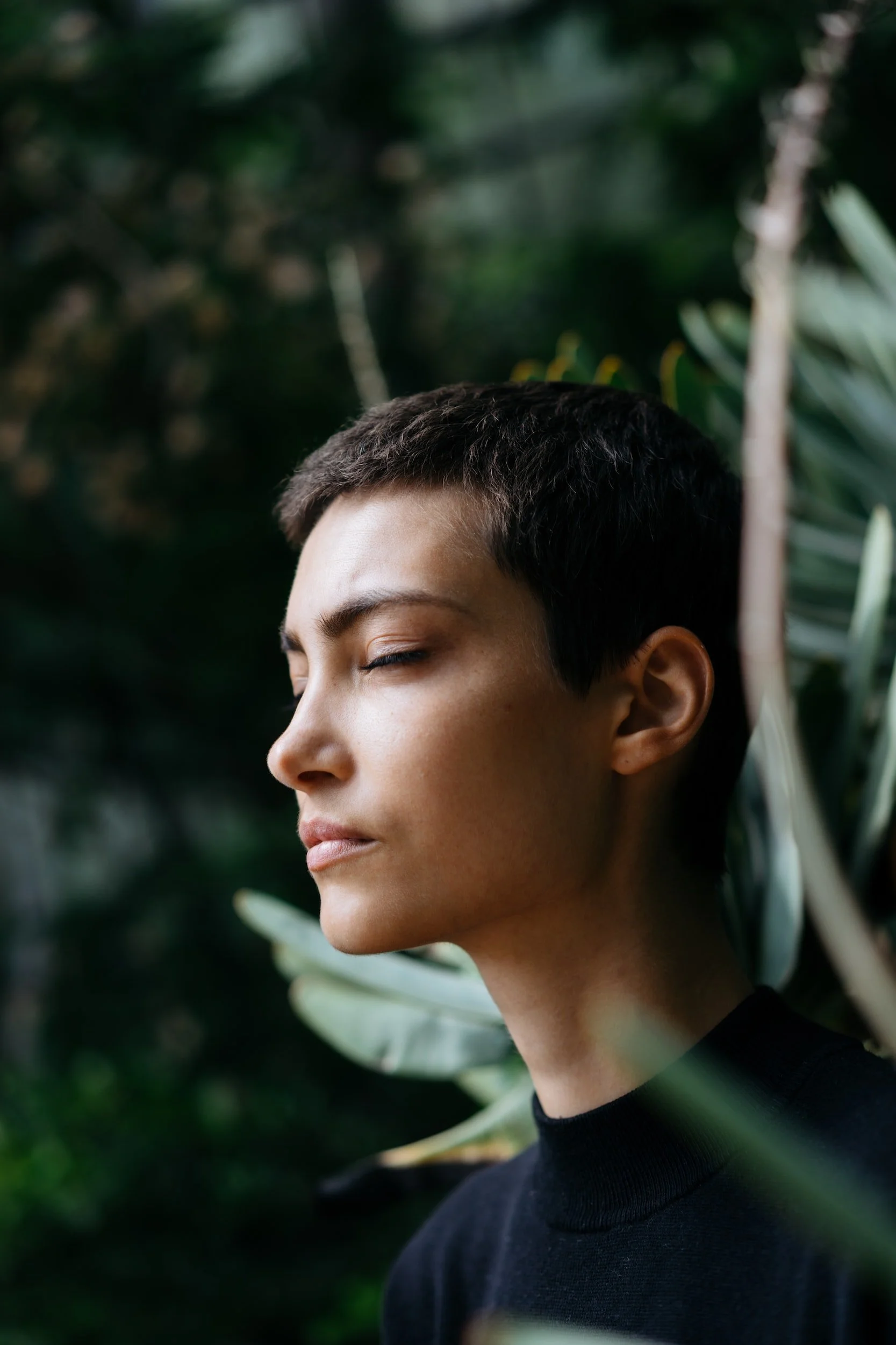 Side profile of young woman with eyes closed, short dark hair, wearing a black top, surrounded by green foliage.