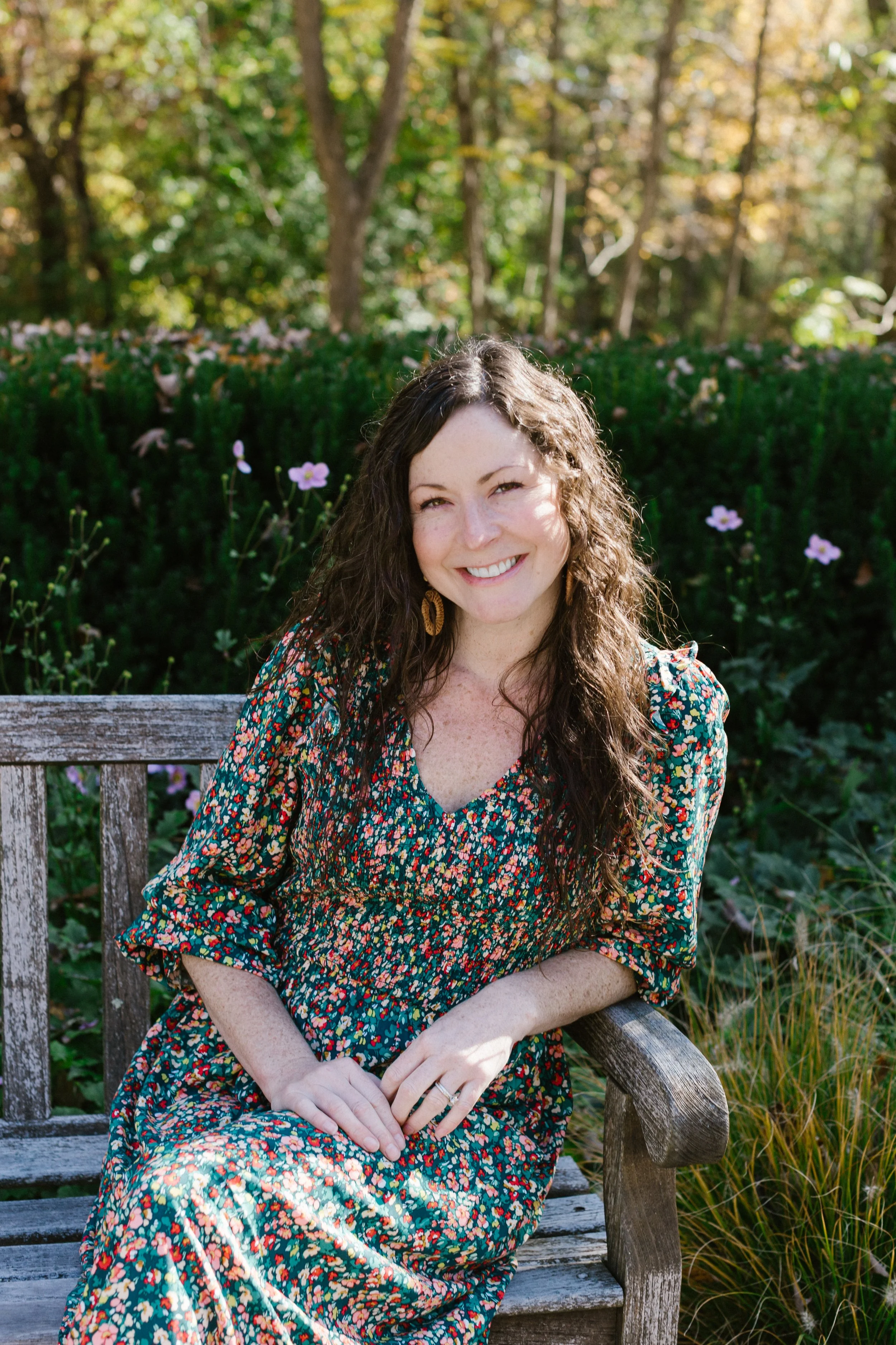 A woman with long curly brown hair is sitting on a wooden bench outdoors, smiling, surrounded by green plants and pink flowers with trees in the background, wearing a colorful floral dress.