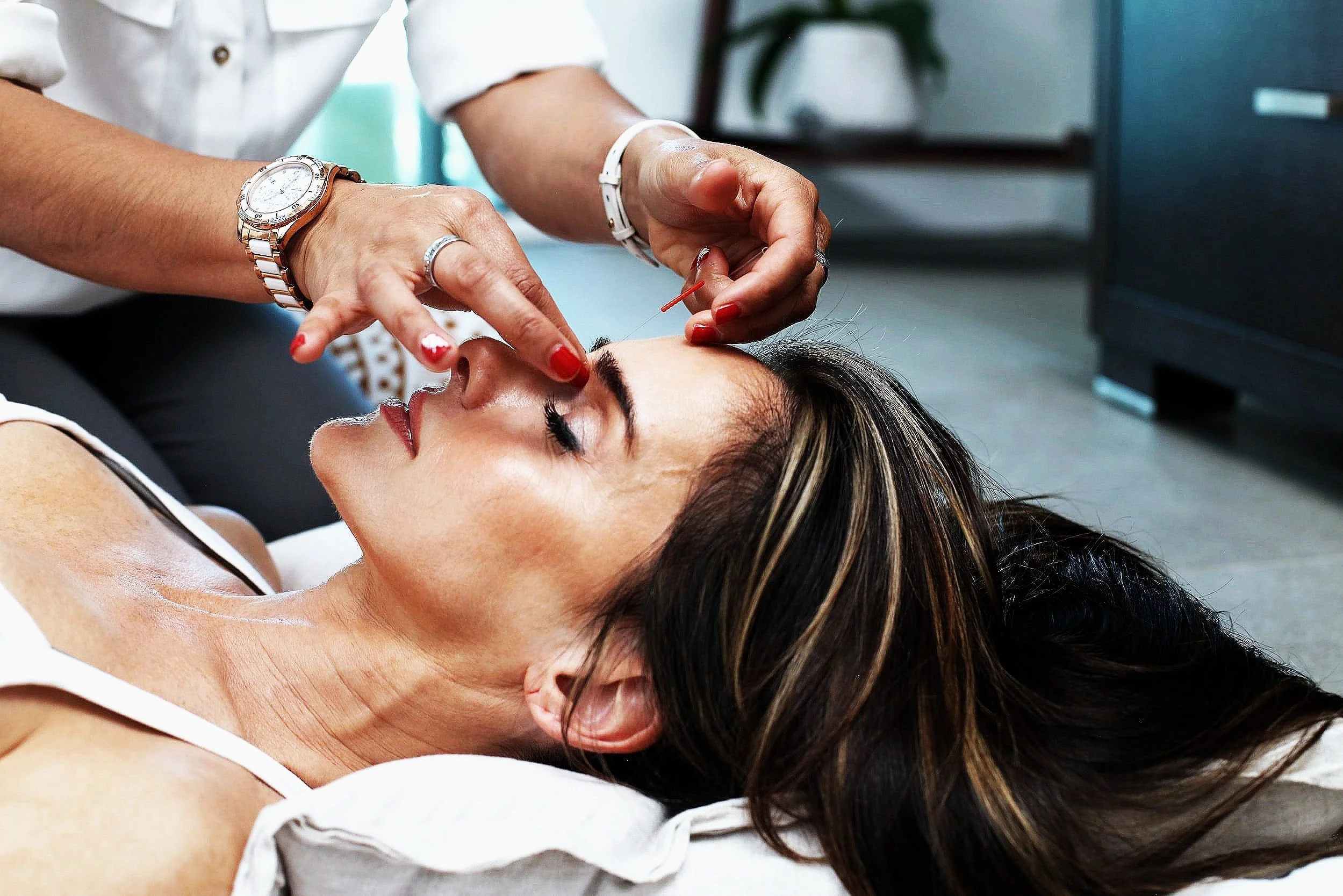 A woman lying down with her eyes closed, receiving a cosmetic injection in her face from a professional healthcare provider.