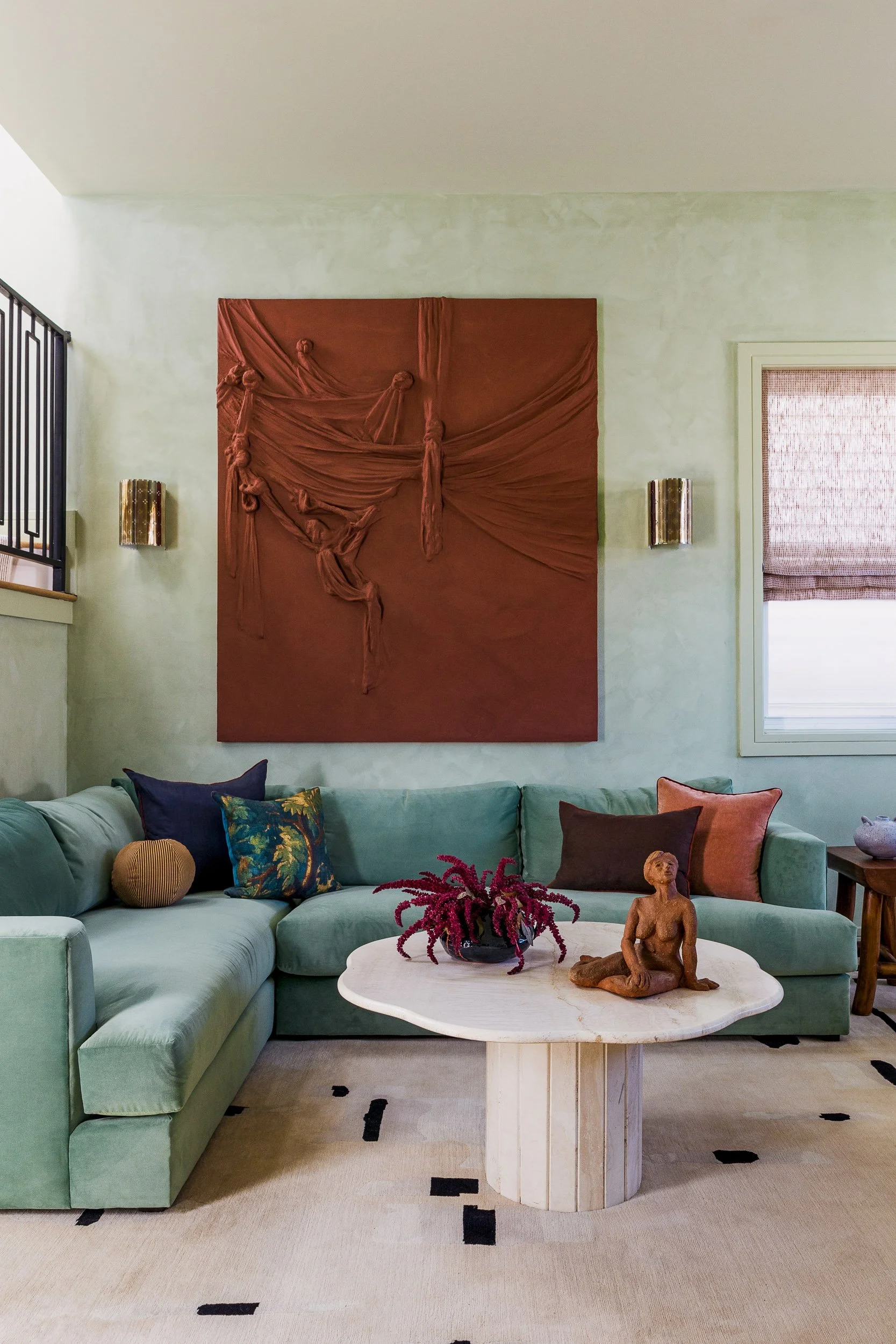 Living room with a mint green sectional sofa, decorative pillows, a white marble coffee table with a sculpture of a seated woman, a plant with pink flowers, a large red textured wall art, and wall-mounted lights.