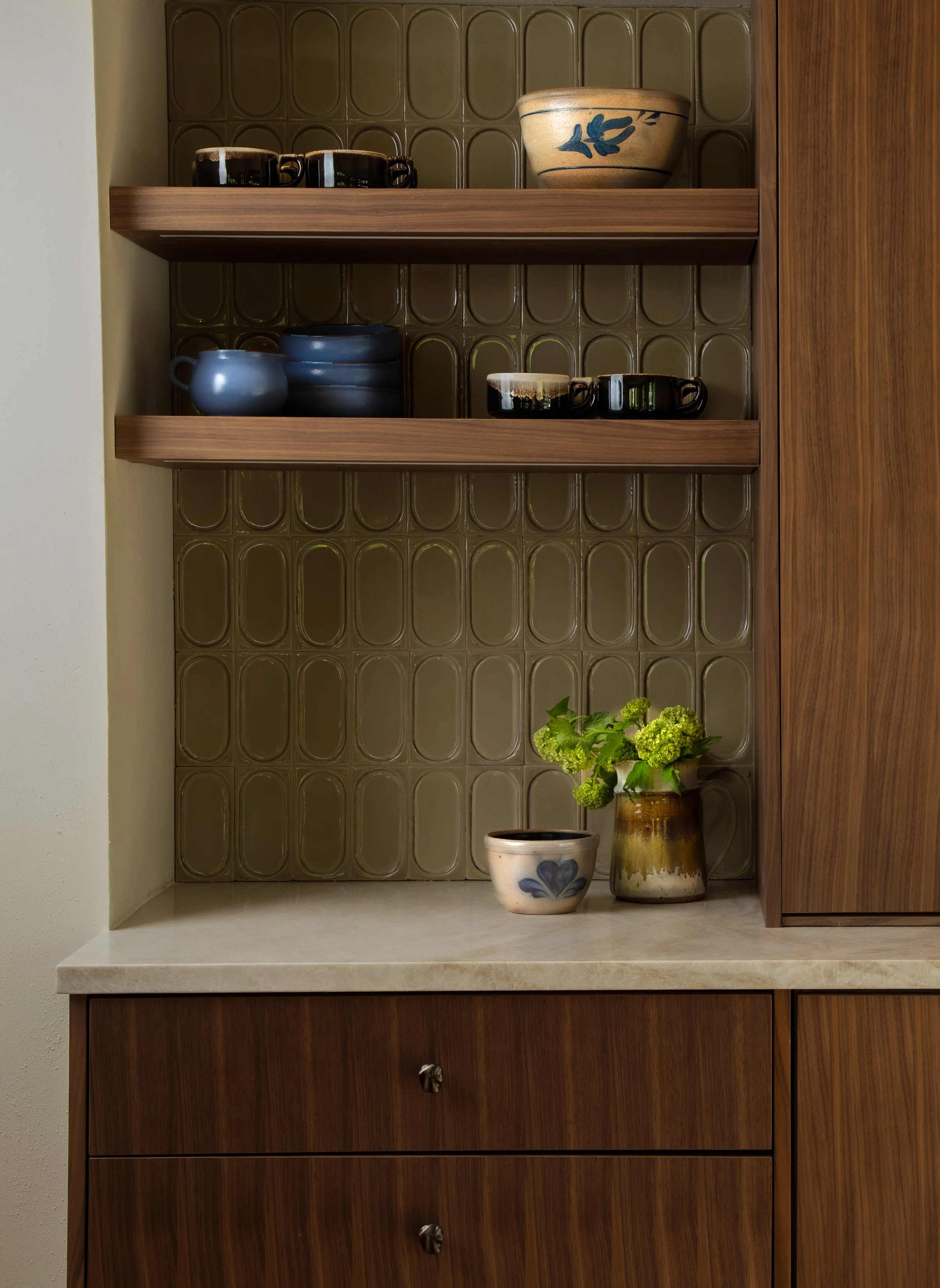 Kitchen shelf with various dishes and ceramics, including bowls, cups, and a vase with green flowers.