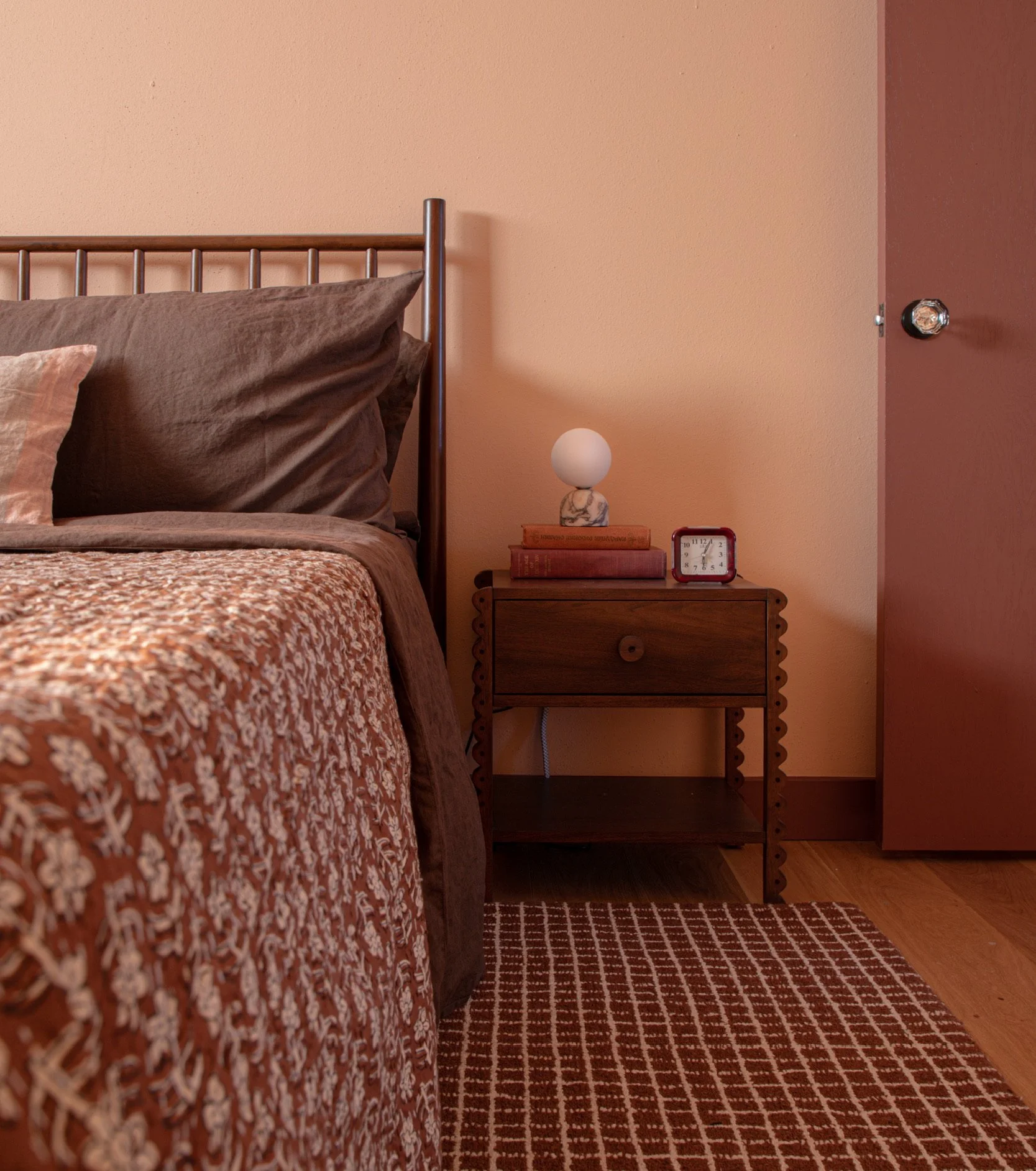 A bedroom corner featuring a bed with patterned bedspread, brown pillows, and a side table with a white spherical lamp, stack of books, a small red clock, against beige and pink walls, and a wooden floor with a reddish-brown patterned rug.