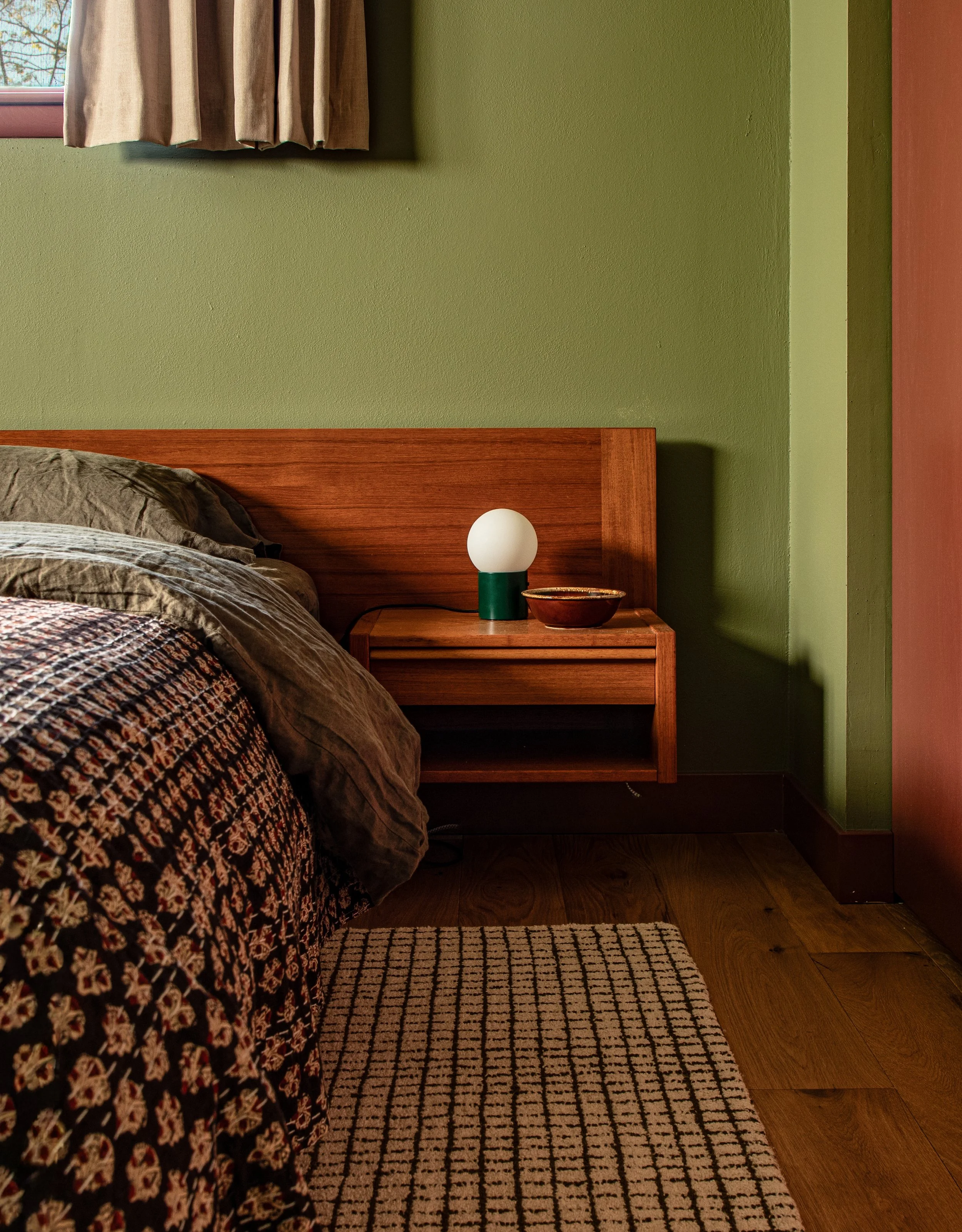 A bedroom corner with a wooden bed, a green wall, a nightstand with a green and white lamp, a bowl, and a patterned rug on brown wooden floor.