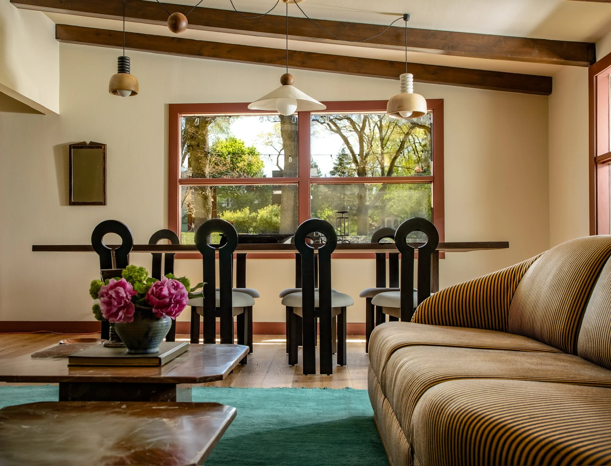 Living room with large window showing trees outside, decorated with modern pendant lights, a striped sofa, a coffee table with flowers, and a green rug.
