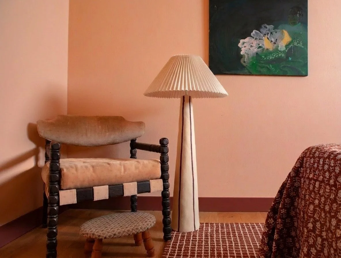 A cozy bedroom corner with peach-colored walls. A vintage wooden armchair with a padded cushion and a small round ottoman are placed to the left. A tall, cream-colored floor lamp with a pleated lampshade is next to the chair. Behind the lamp, a dark 