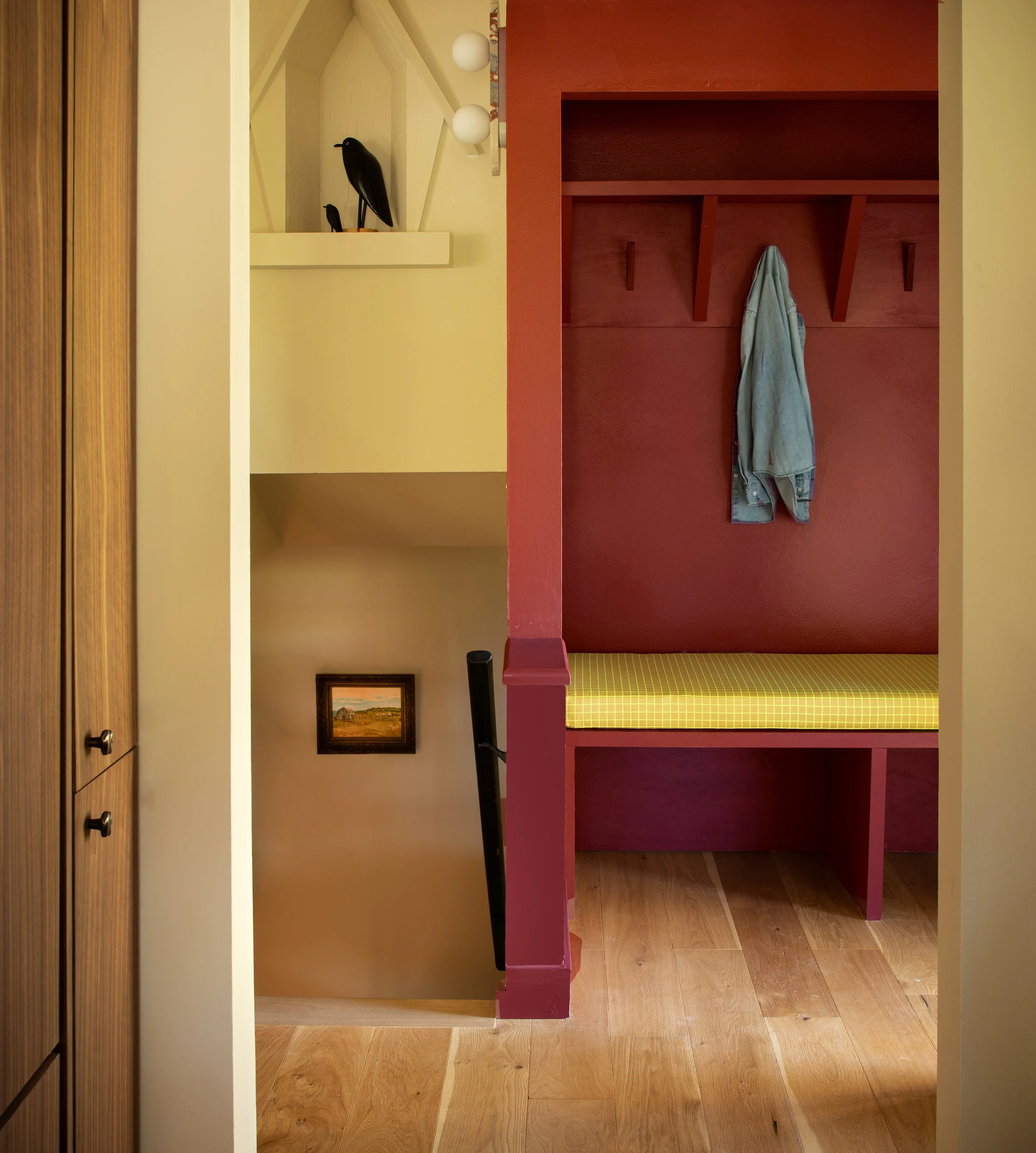 Interior view of a cozy room with a wooden wardrobe on the left, a red wall with a built-in coat hook and a hanging denim jacket, a photo of a rural landscape on the wall, and a corner with yellow patterned seating. The floor is wooden.