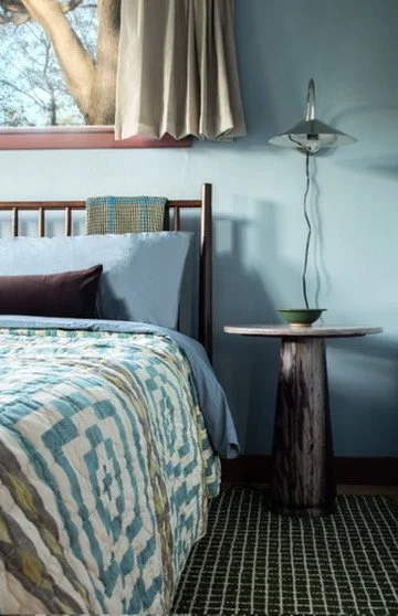 A bedroom corner with a bed, pillow, and a side table with a bowl, next to a wall with a wall-mounted lamp and a window with beige curtains.
