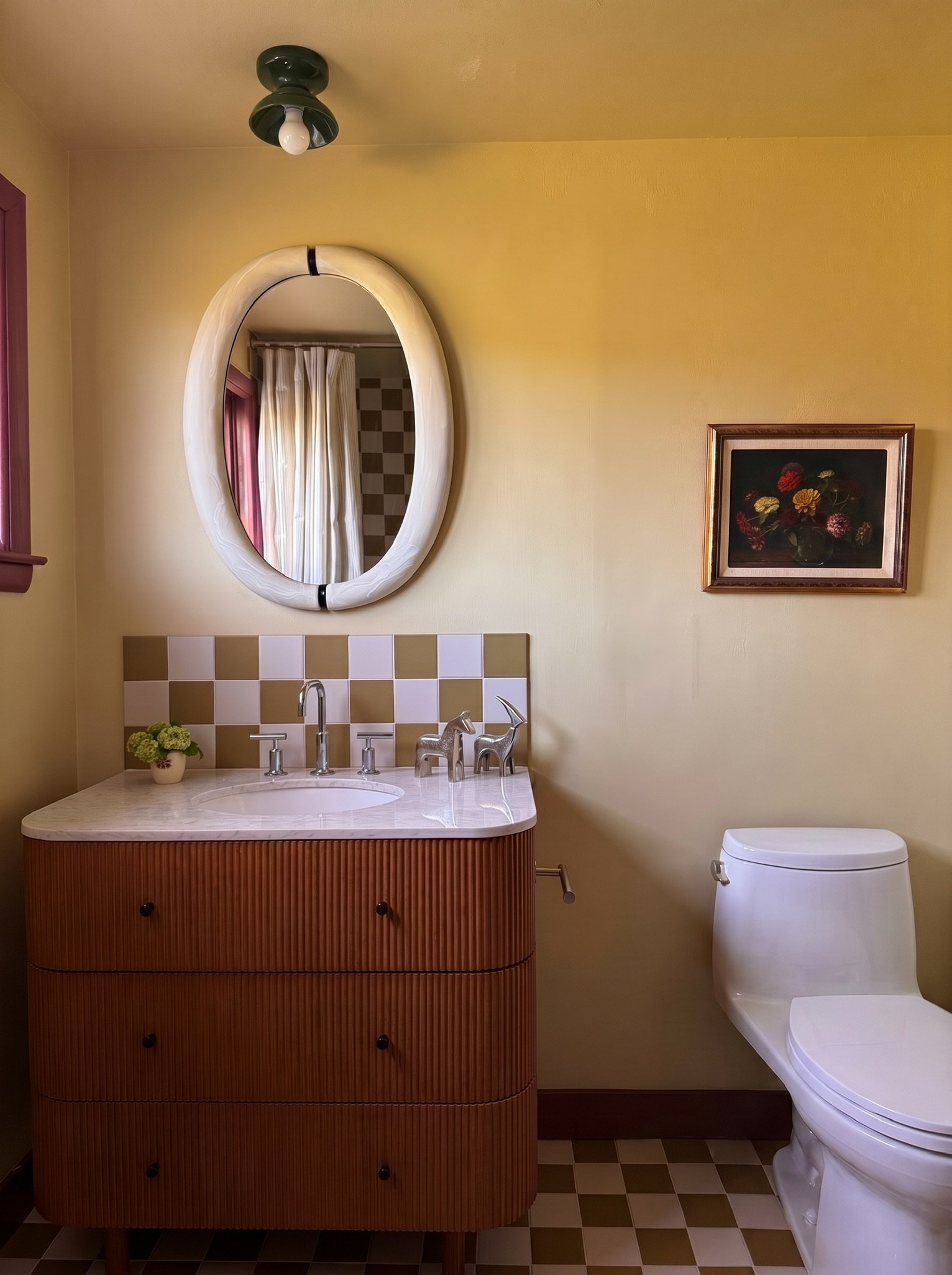 A bathroom with a yellow wall, a white oval mirror, a wooden vanity with a sink, and a white toilet. Framed floral artwork hangs on the wall, and a black ceiling light fixture is visible. The vanity has a potted plant and a decorative dog figurine.