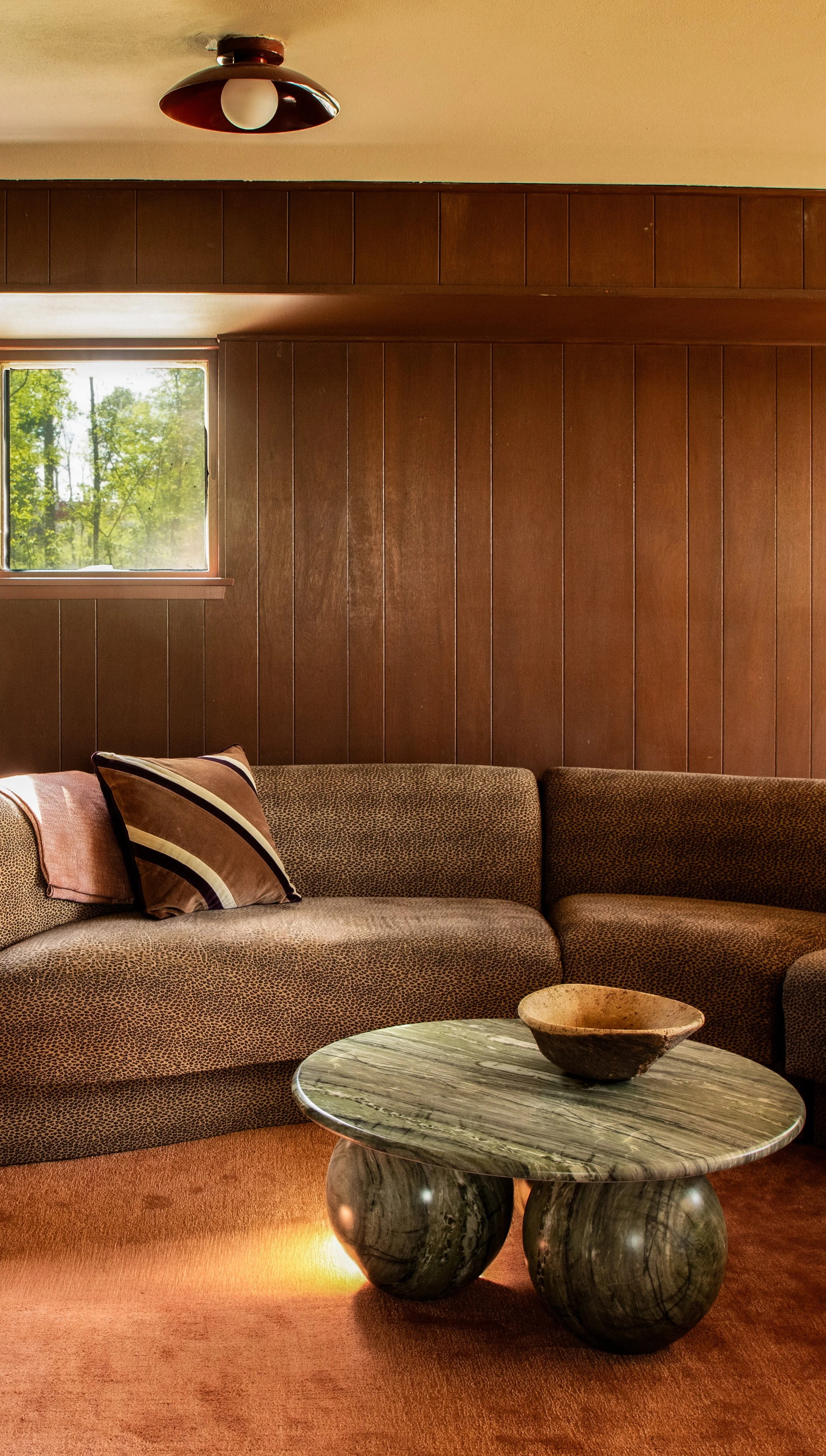 A living room with a brown patterned sectional couch, a throw pillow, a round marble coffee table with a bowl, a wood-paneled wall, a small window with a view of trees, and a ceiling light fixture.