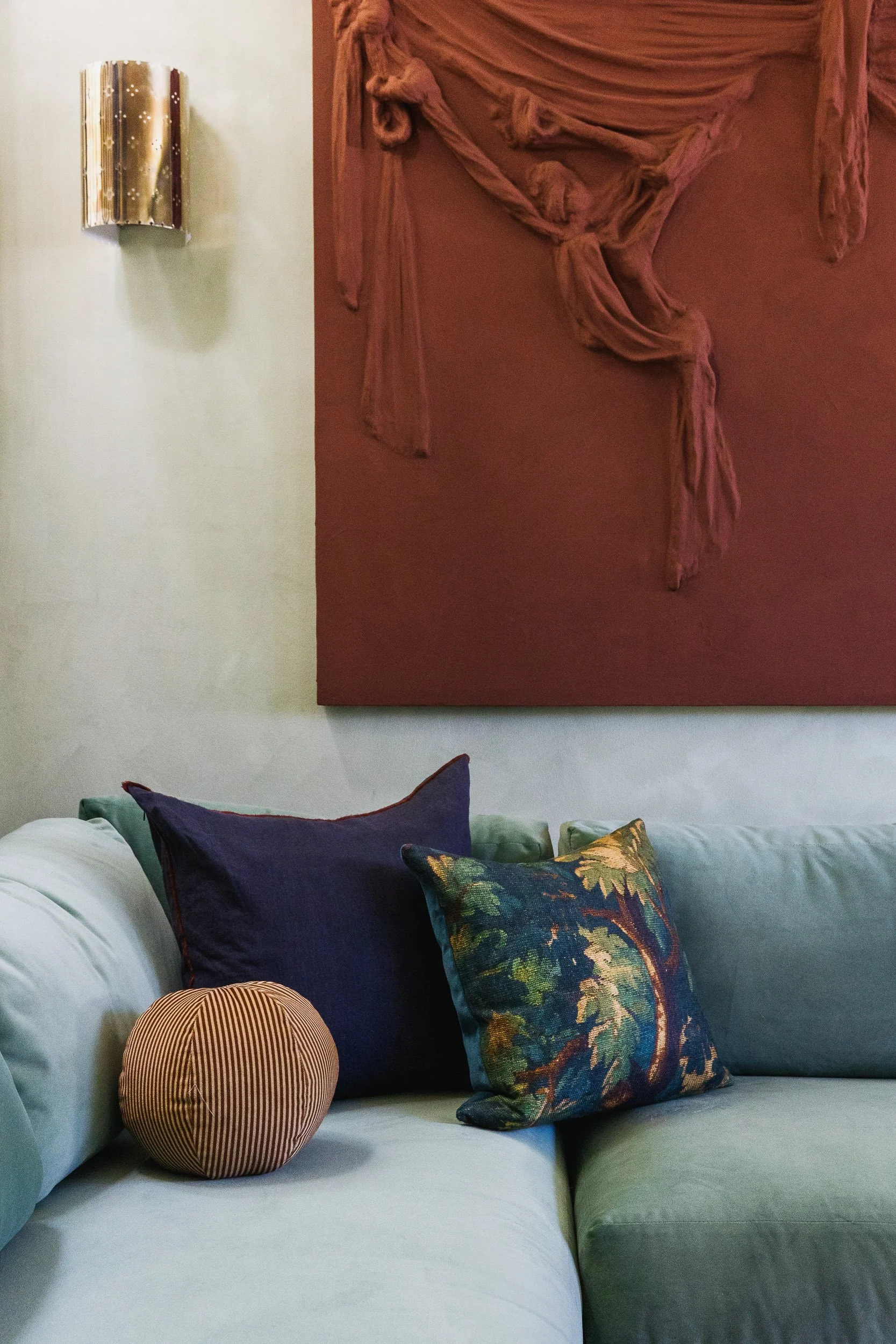 Living room scene with a light-colored sofa decorated with multiple pillows, including purple, floral, and striped patterns. A round striped pouf is placed on the sofa, and a large abstract painting with draped fabric in shades of pink is hung on the wall behind the sofa.