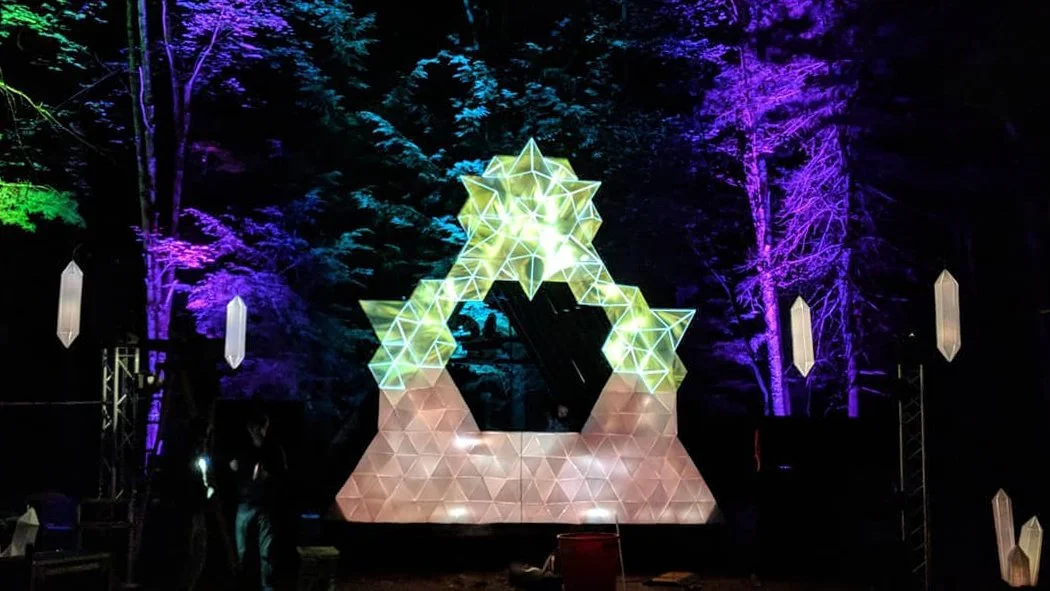 Photo of the Crystal Forest installation.  A festival stage design featuring projection-mapped geometric shapes and colorful ambient lighting on trees.