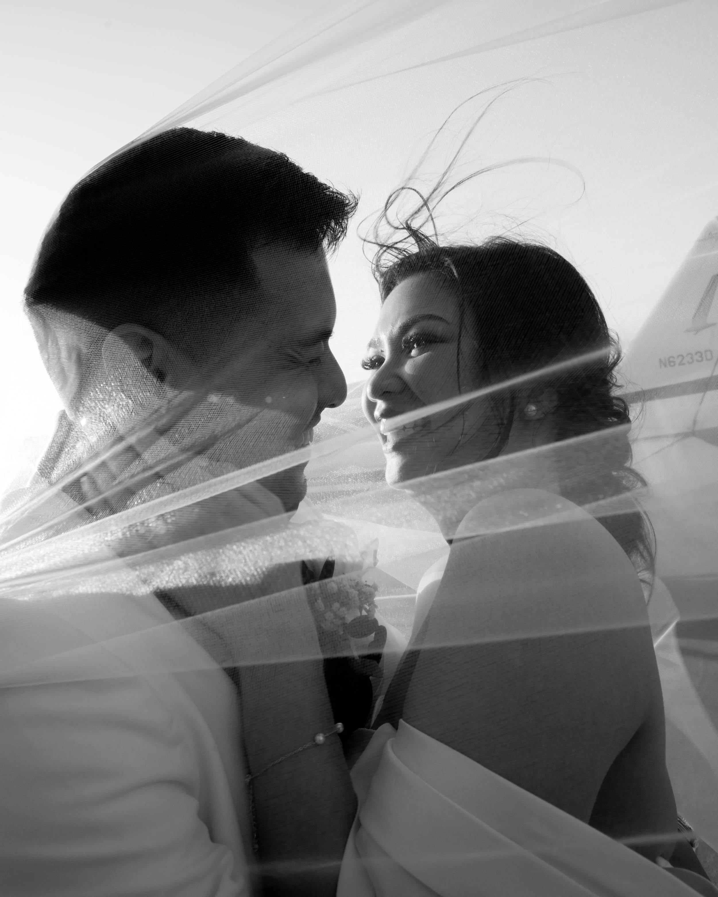 A couple in wedding attire embracing in front of small airplanes on an airport tarmac.
