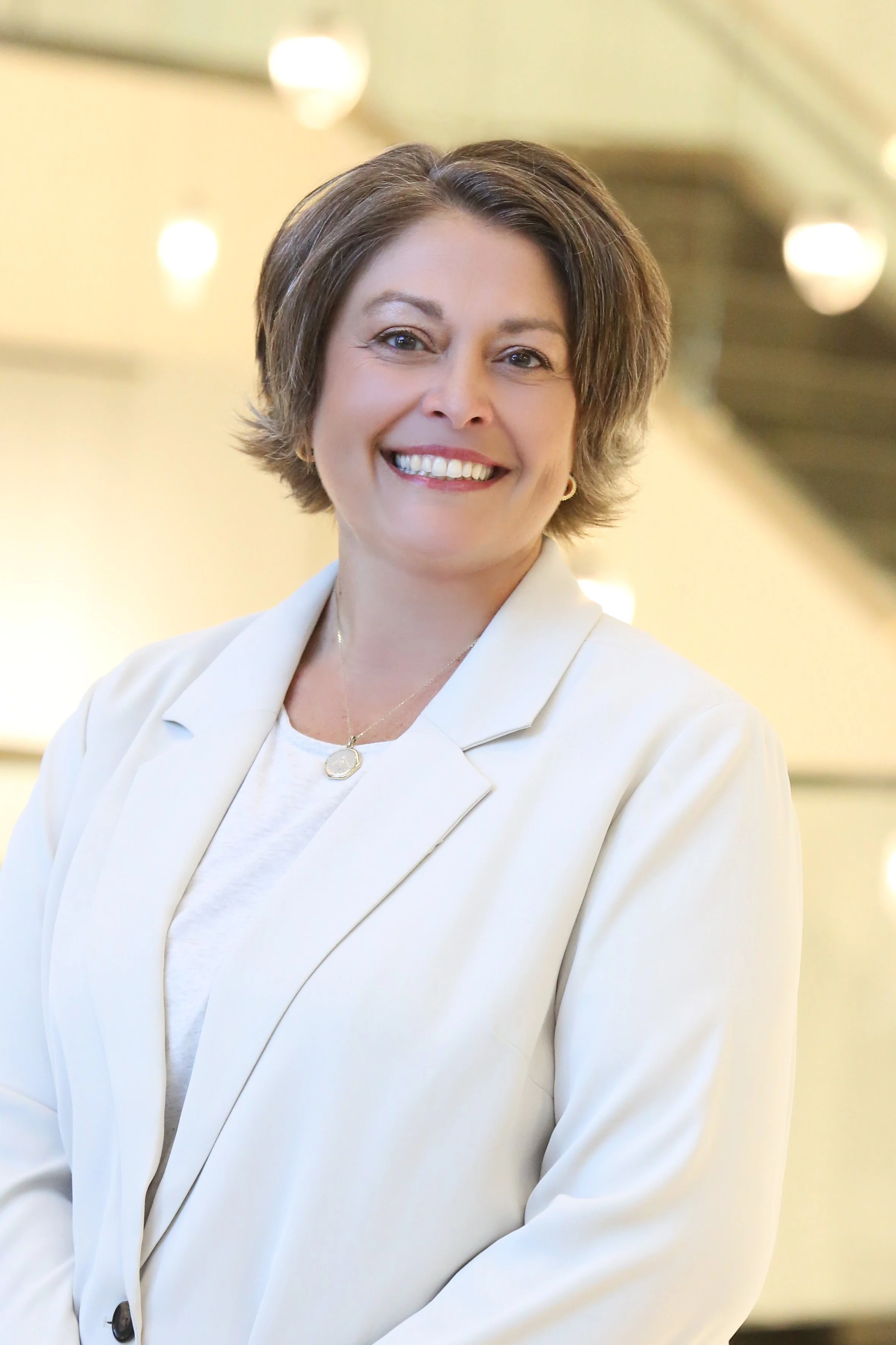 A smiling woman with short brown hair wearing a cream-colored blazer and a gold necklace, standing indoors with blurred circular lights in the background.