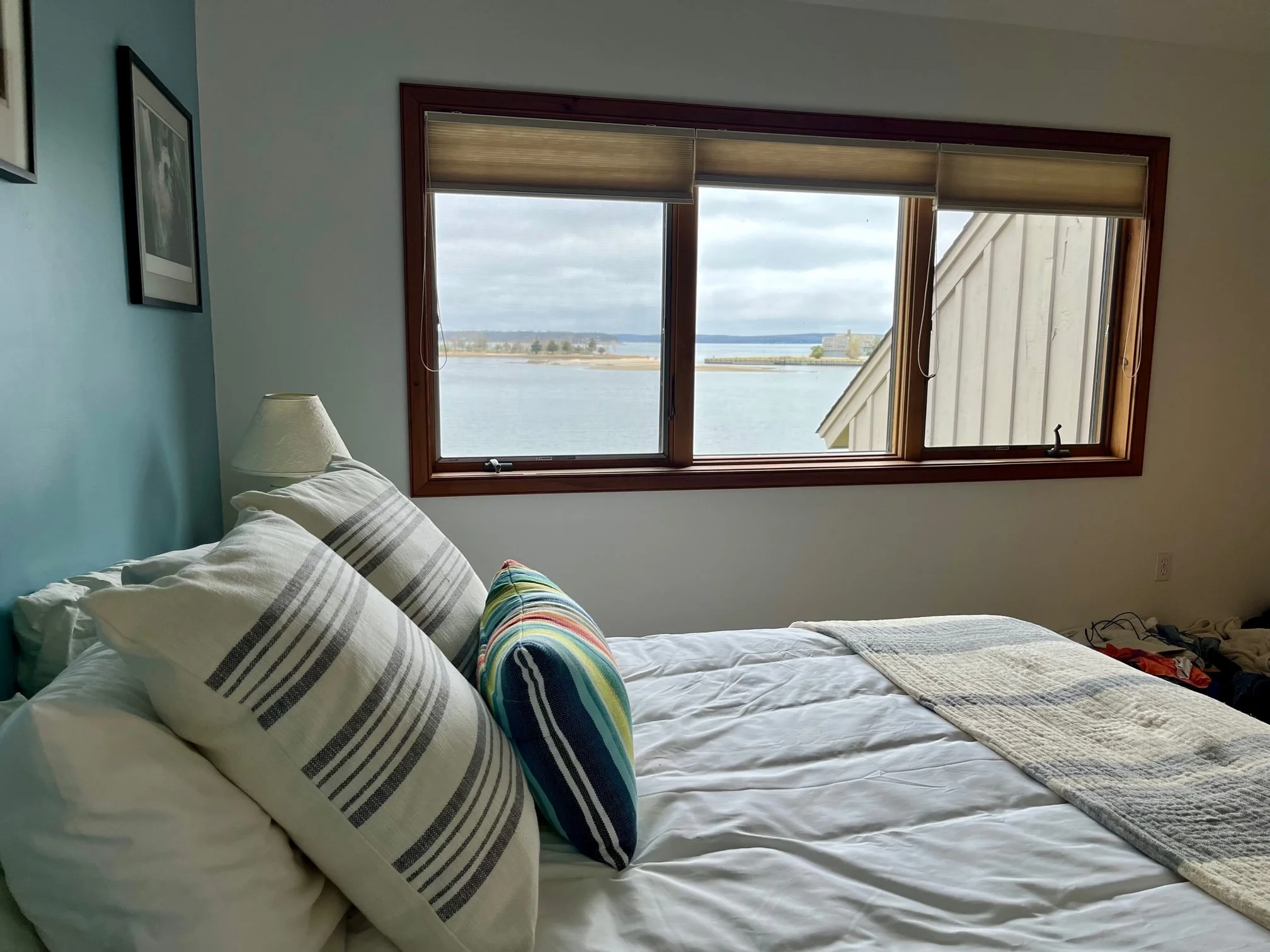 A cozy bedroom with a view of a body of water through a large window. The bed has white sheets, striped pillows, and a colorful oval pillow. There is a lamp beside the bed and framed pictures on the blue wall.