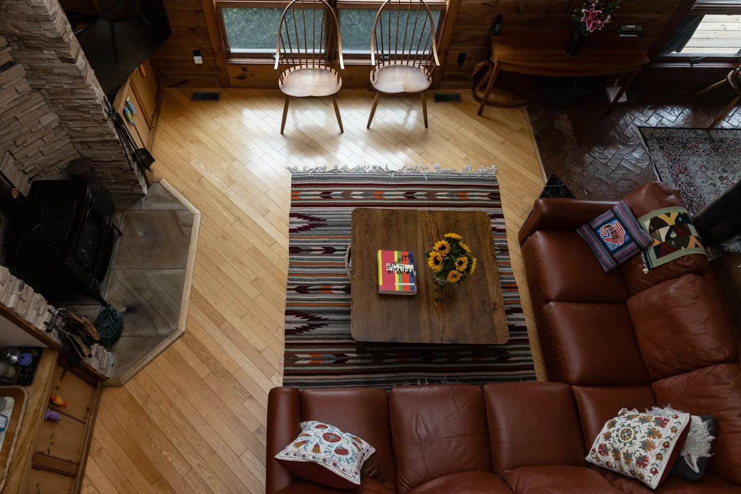 Living room with hardwood floors, a stone fireplace, a wooden coffee table with sunflowers and colorful books, an L-shaped leather sofa with decorative pillows, two wooden chairs near a large window, and a corner desk with a potted plant.