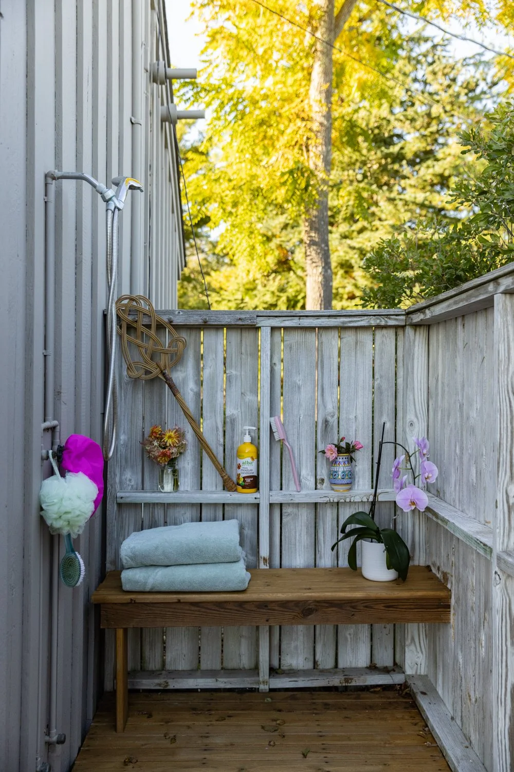 An outdoor shower area with a wooden bench, towels, toiletries, and potted plants on a wooden fence.