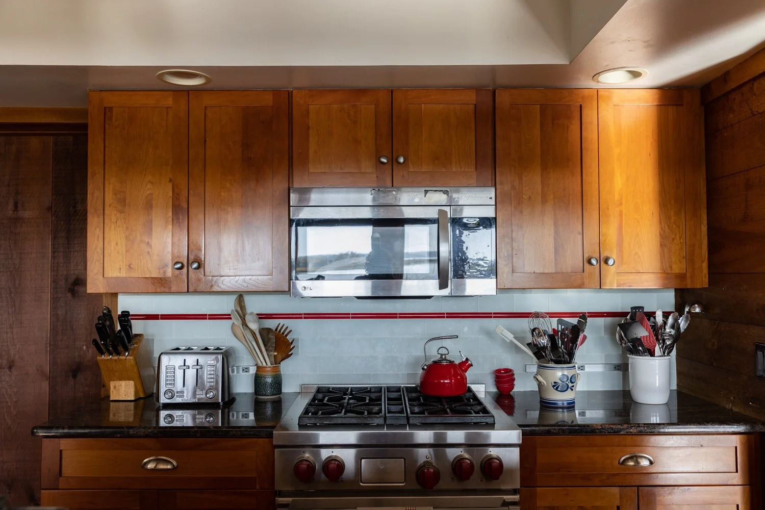 Kitchen with wooden cabinets, stainless steel microwave, gas stove with red kettle, and various utensils and appliances on dark countertop