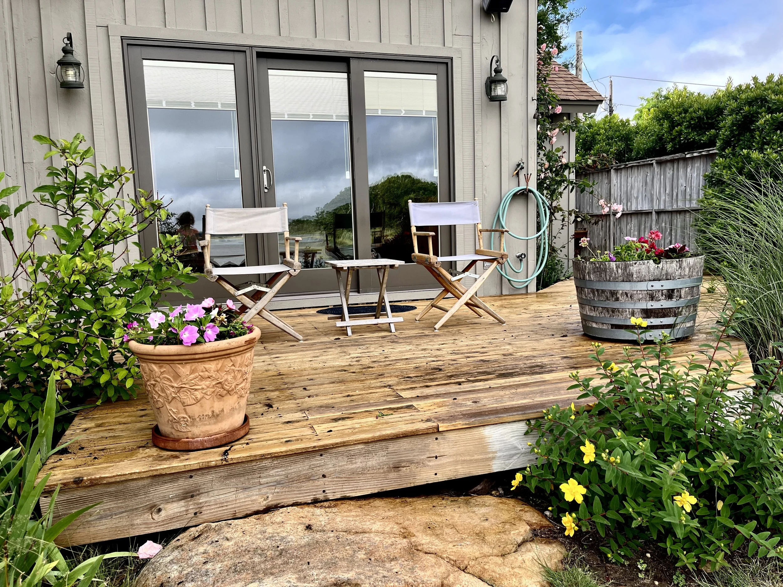 A wooden deck with two white and wood foldable chairs, a small table, potted flowers, garden hose, front of a house with a glass sliding door, surrounding greenery, and cloudy sky.