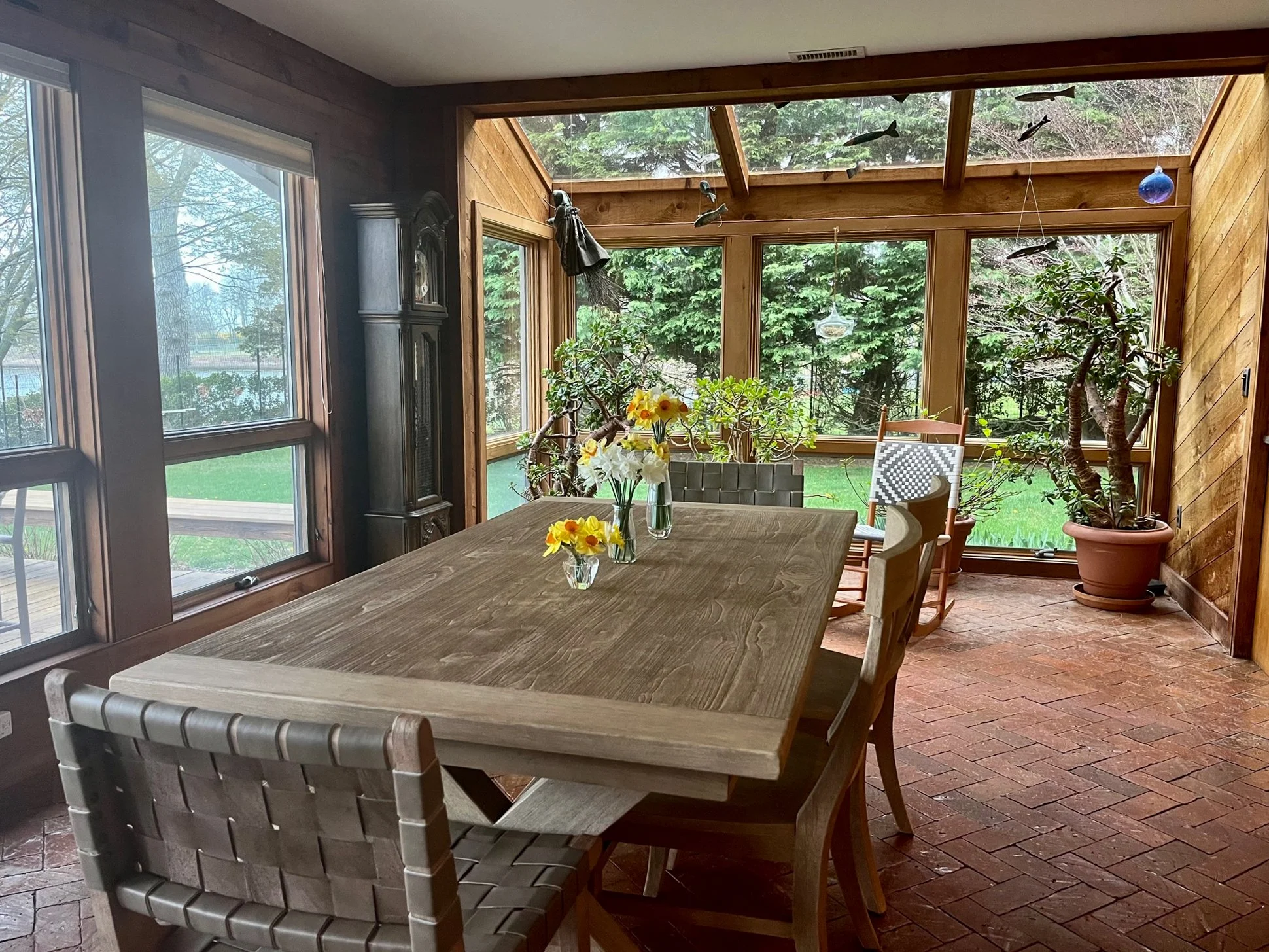 Sunroom with large windows, a wooden dining table with flowers, potted plants, and hanging decorations of fish, surrounded by greenery outside.